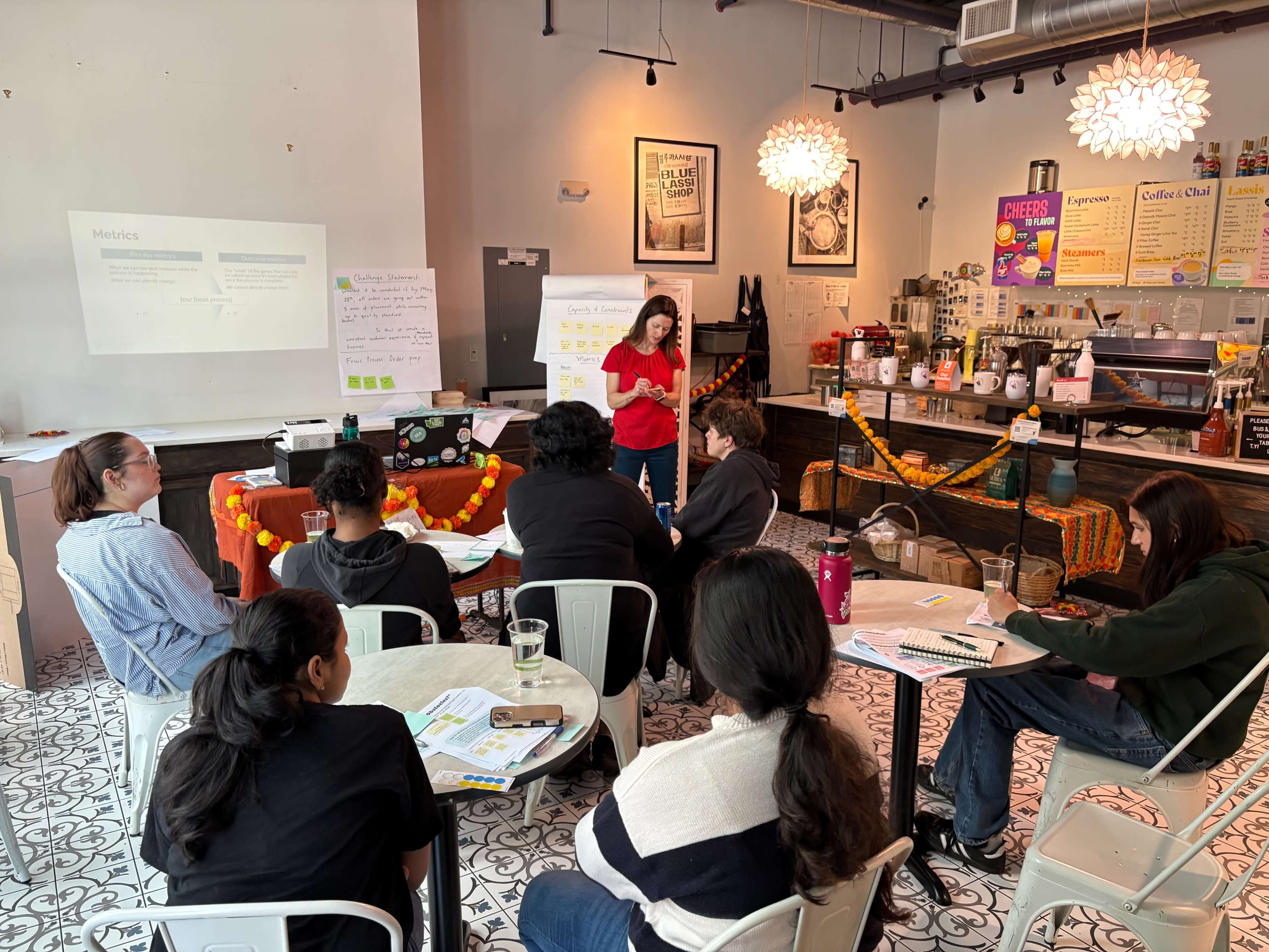 A group of people is seated in a café, attentively listening to a speaker presenting information on a projector while materials are displayed on a table.