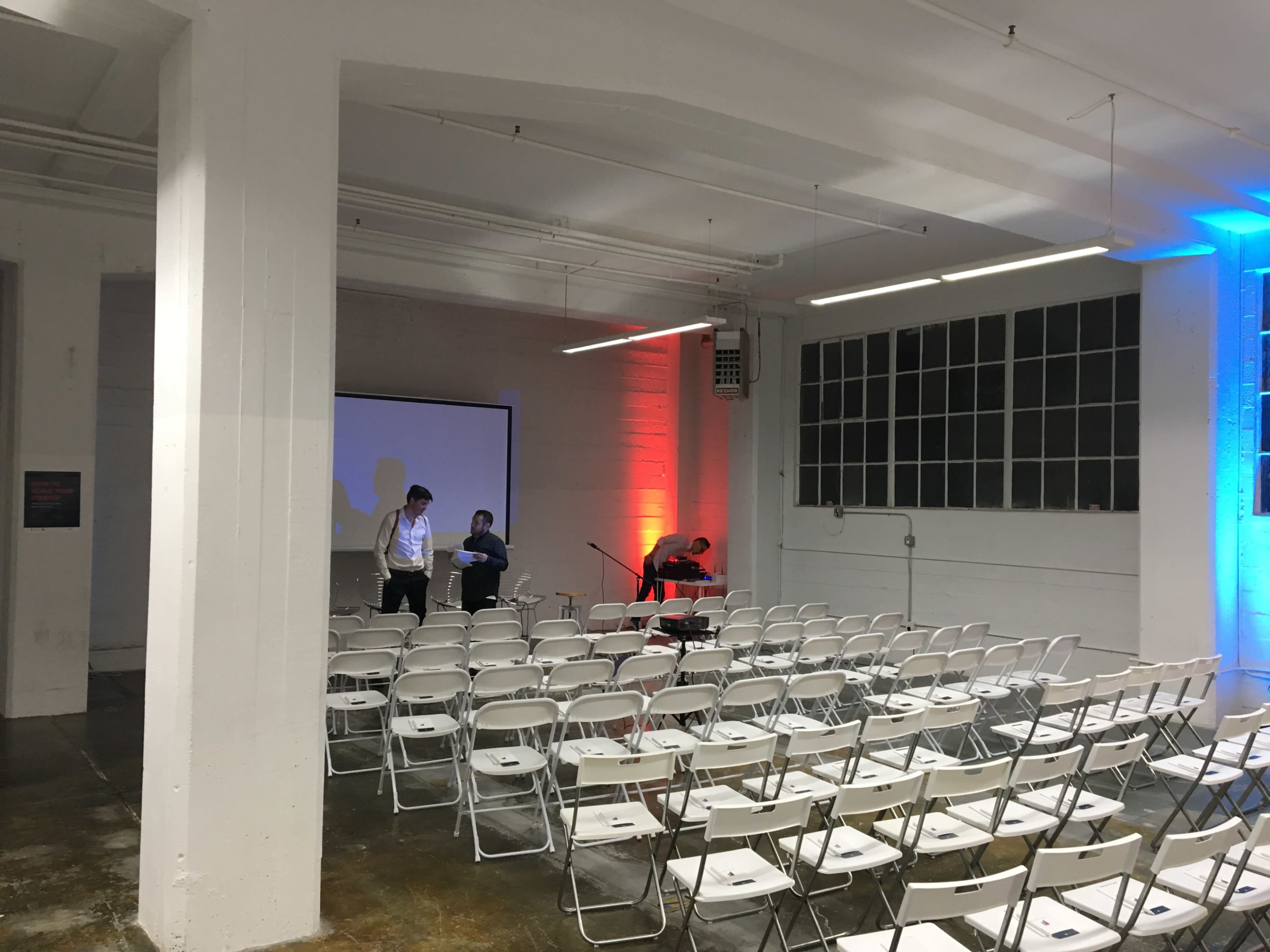 A spacious room with rows of white folding chairs set up for an event, and two people preparing a presentation on a stage with a large screen.