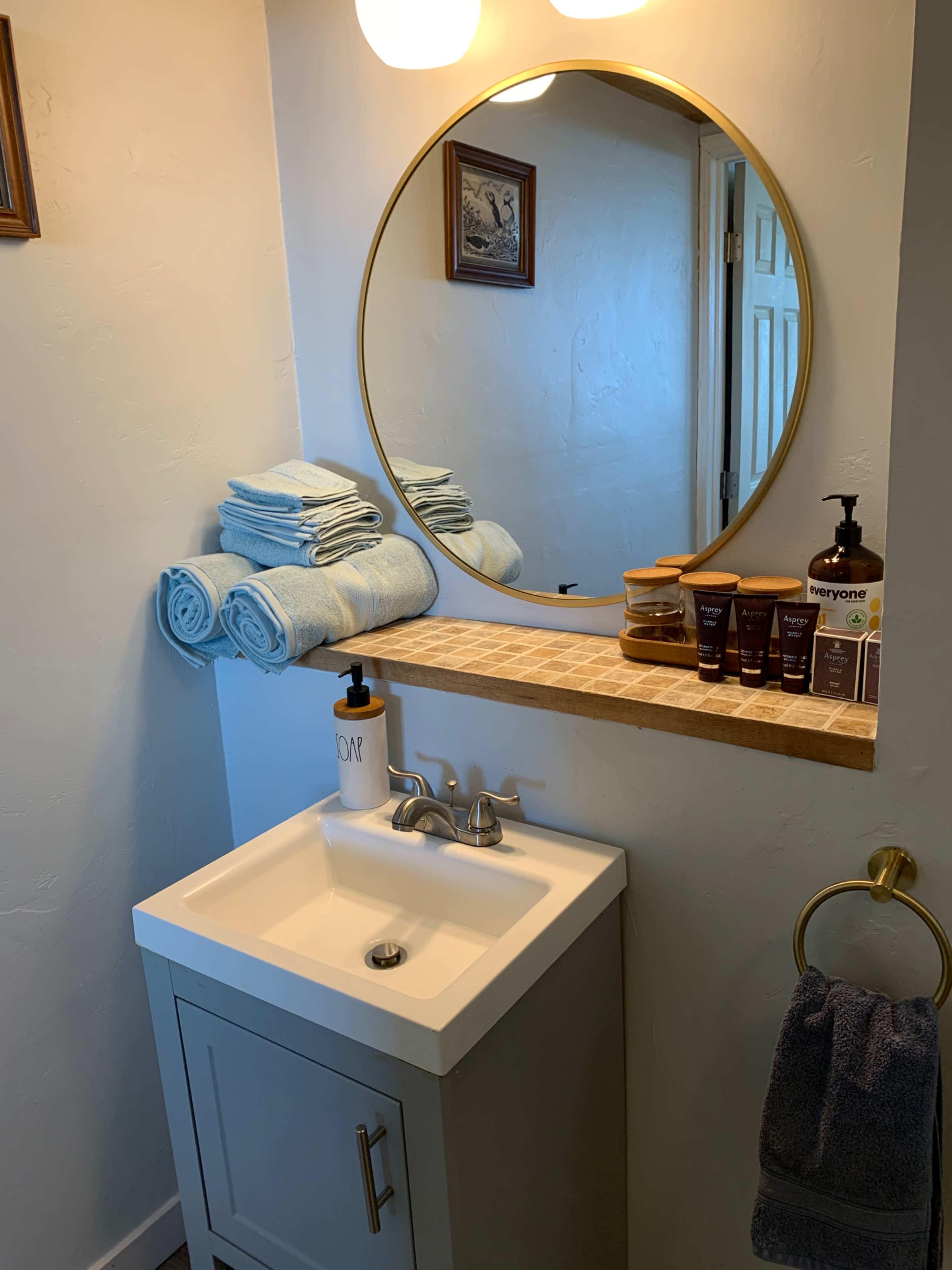 A compact bathroom features a circular mirror above a sink with a modern vanity, neatly arranged towels, and a shelf displaying skincare products.