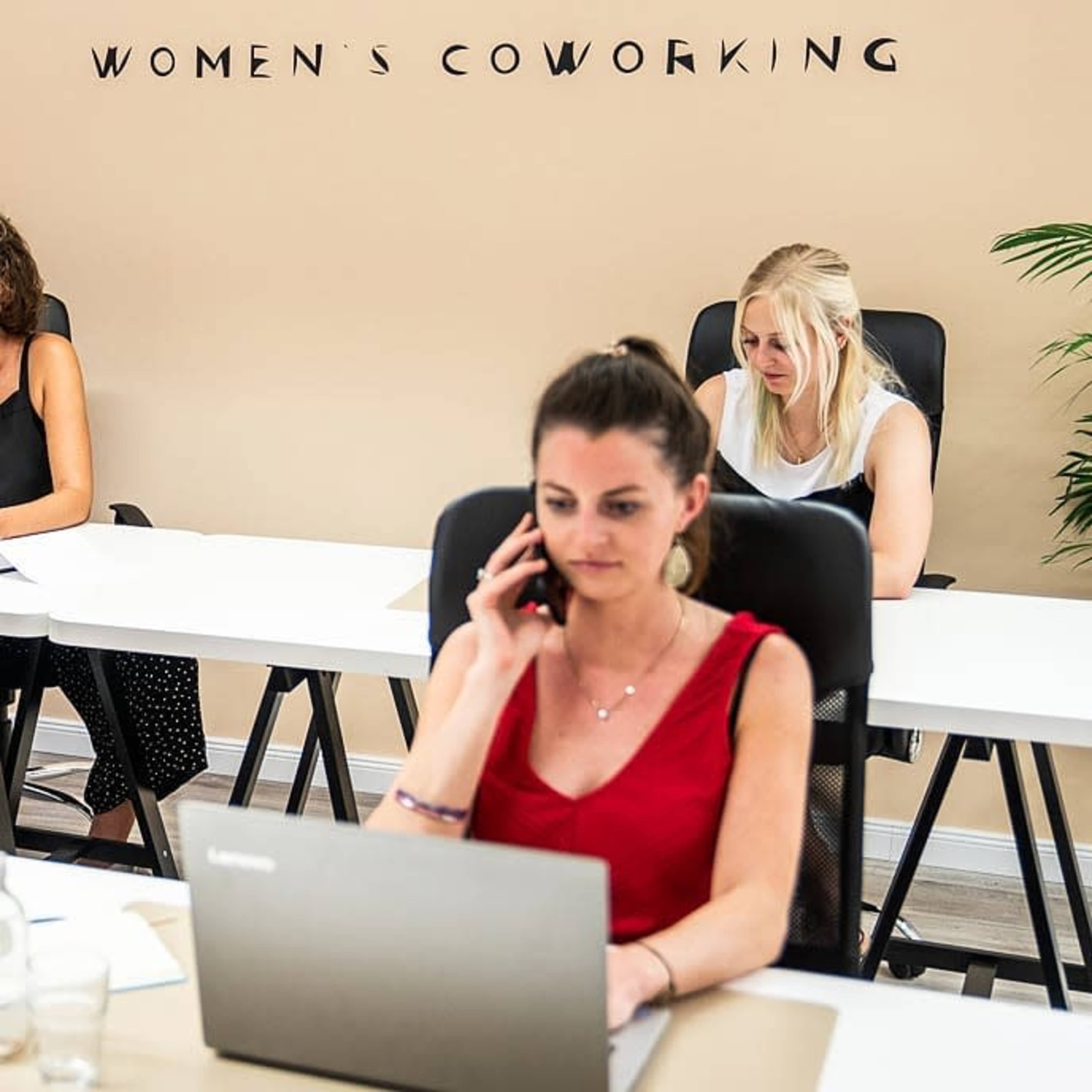 A women's coworking space with three women working at desks, one on the phone and two focused on their laptops.
