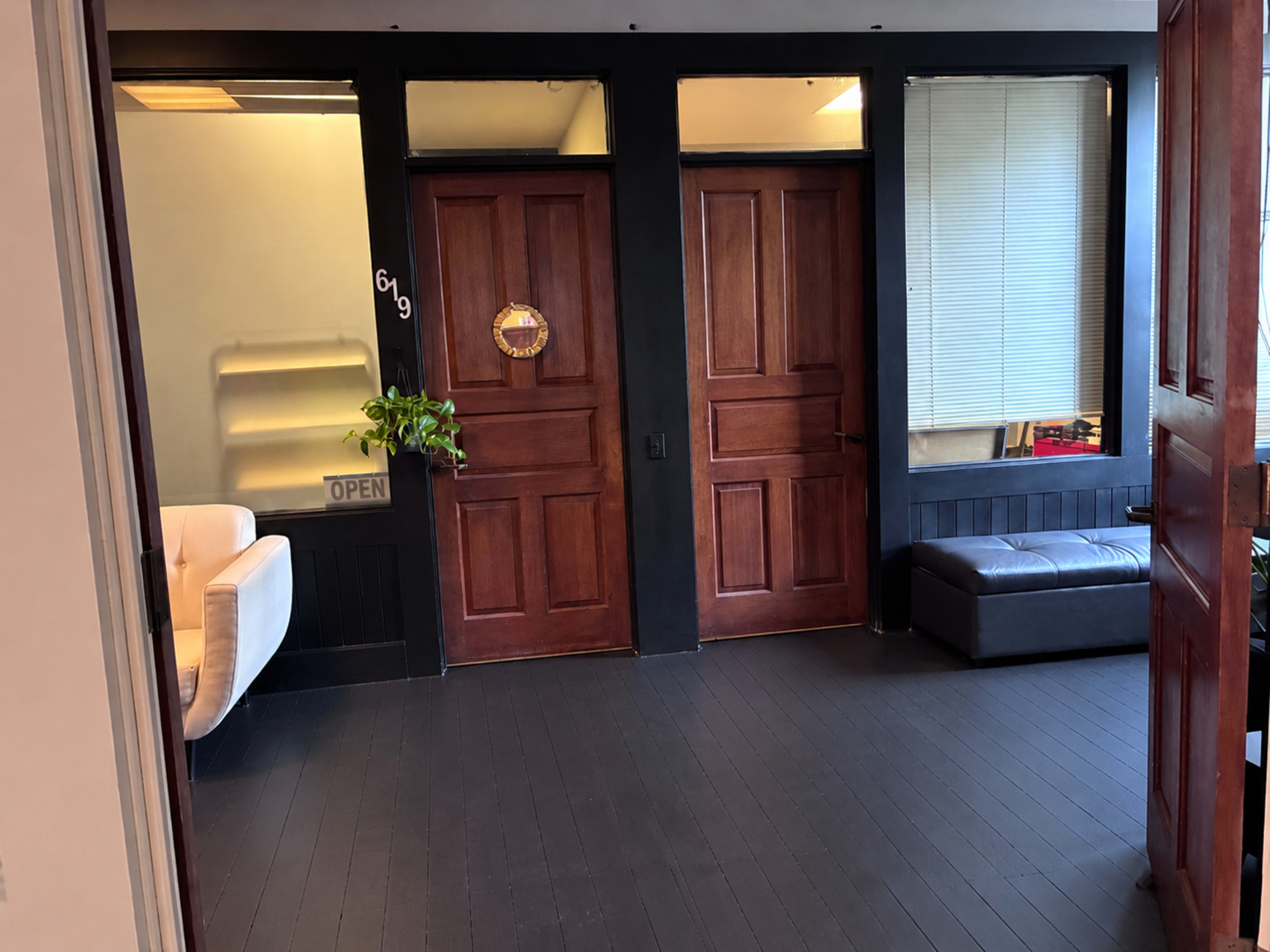 The image shows an interior hallway with two wooden doors, a small green plant on a ledge, a couch, and a sign that reads "OPEN."