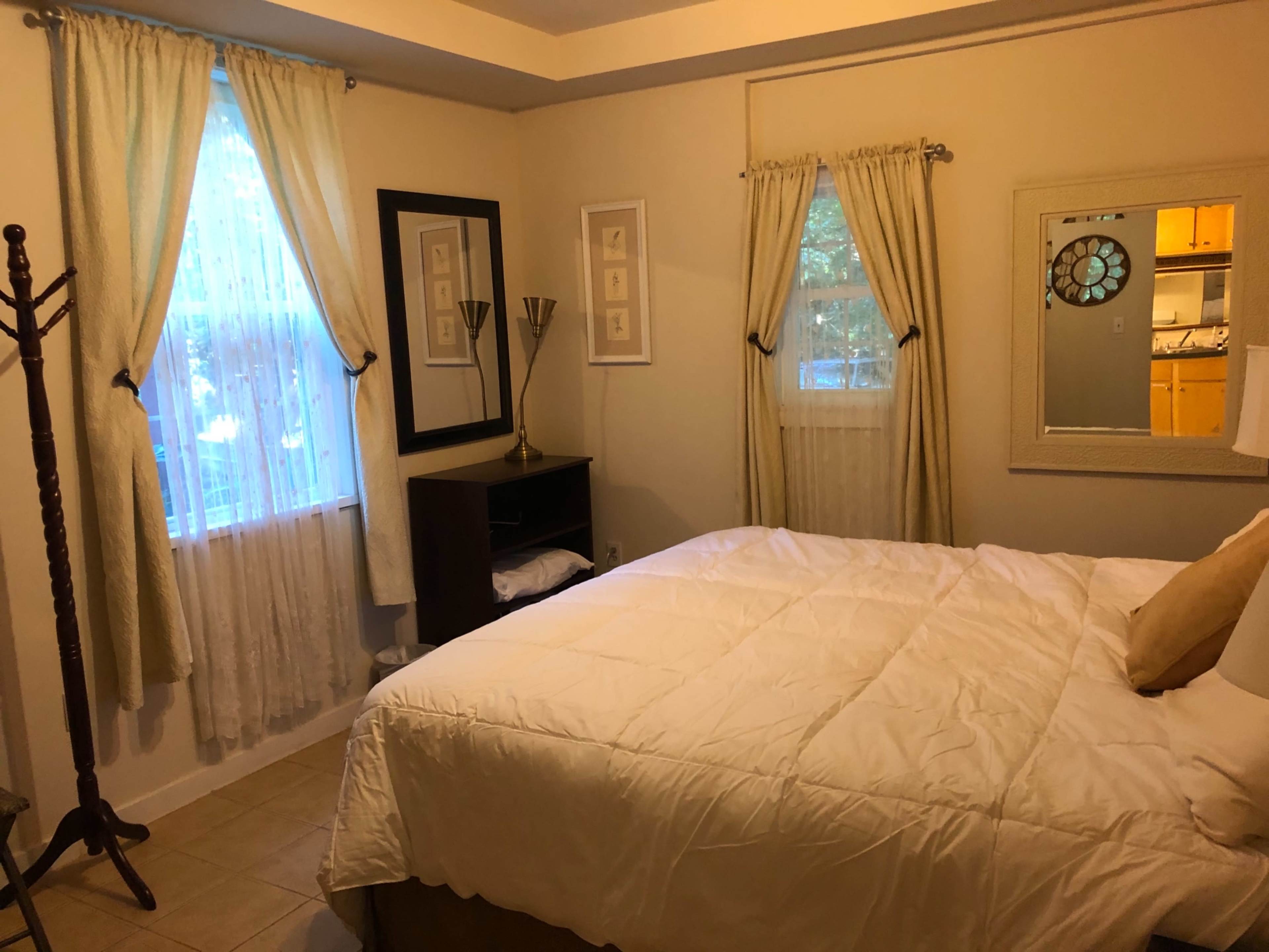 The image shows a bedroom with a large bed, light-colored curtains, and a mirror on the wall, beside a window that lets in natural light.