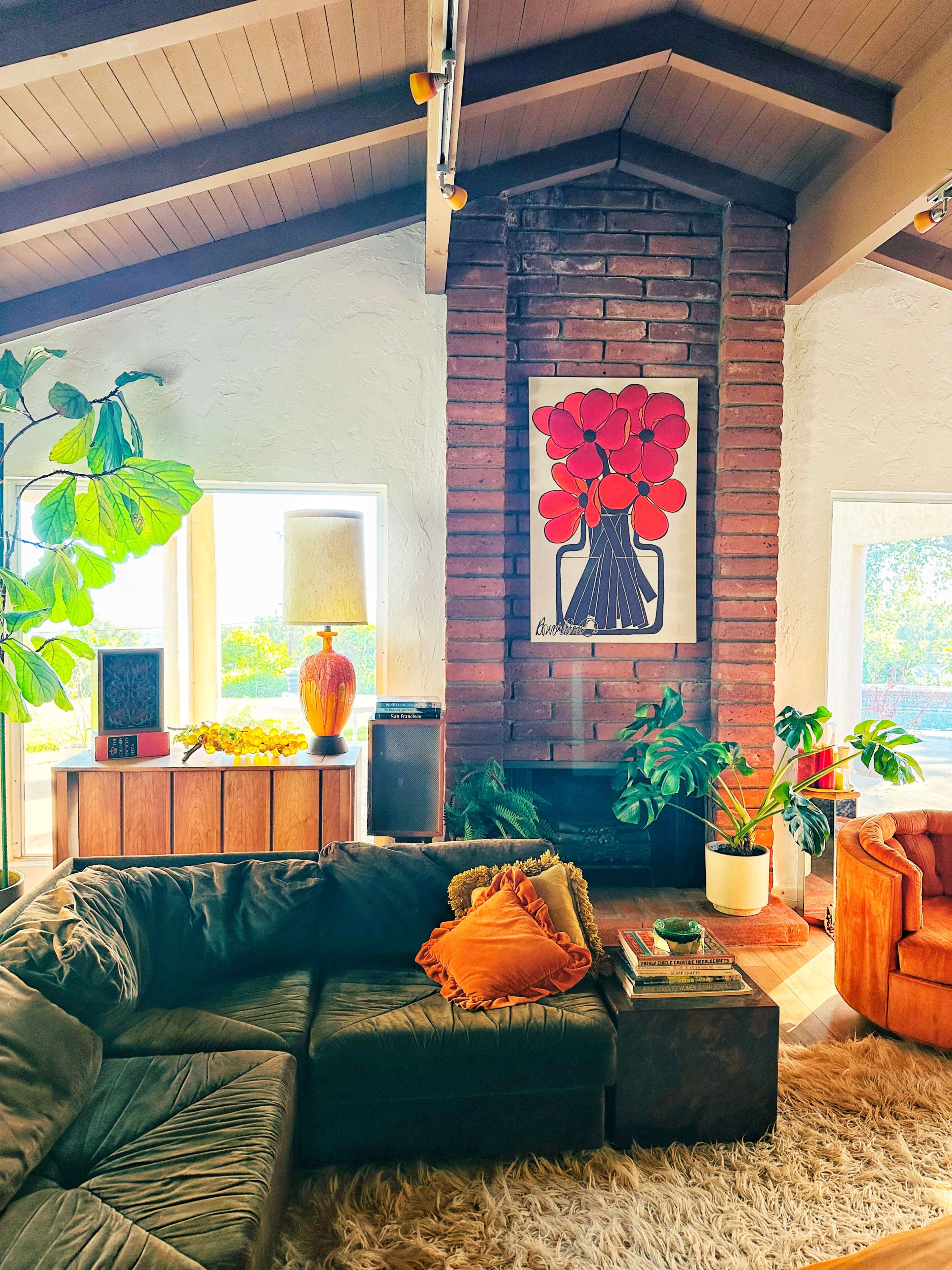 The image shows a cozy living room featuring a green sectional couch, an orange armchair, a brick fireplace with a floral artwork above it, and various plants and decor items.