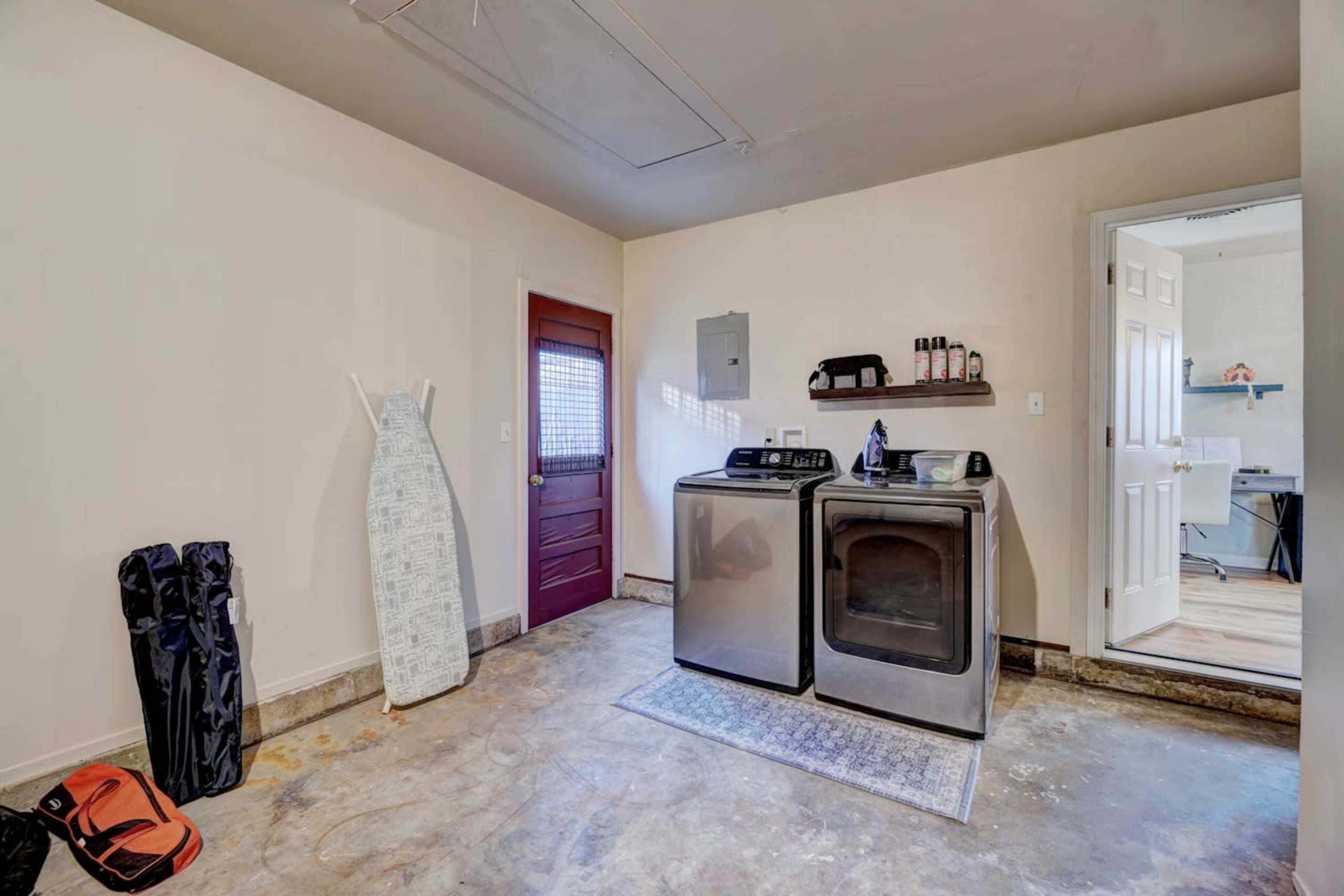 A laundry room with a washer and dryer, an ironing board, and a red door leading to another room.