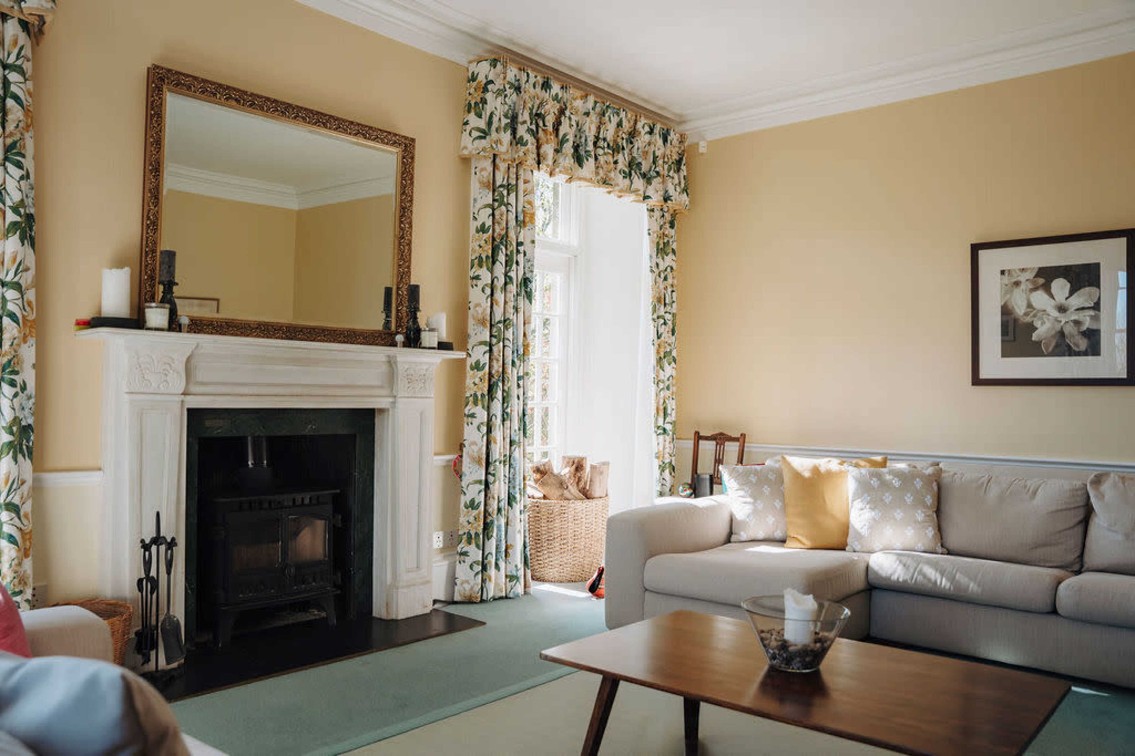 The image shows a living room with a fireplace, a large mirror, and floral curtains, featuring a light-colored sofa and a wooden coffee table.