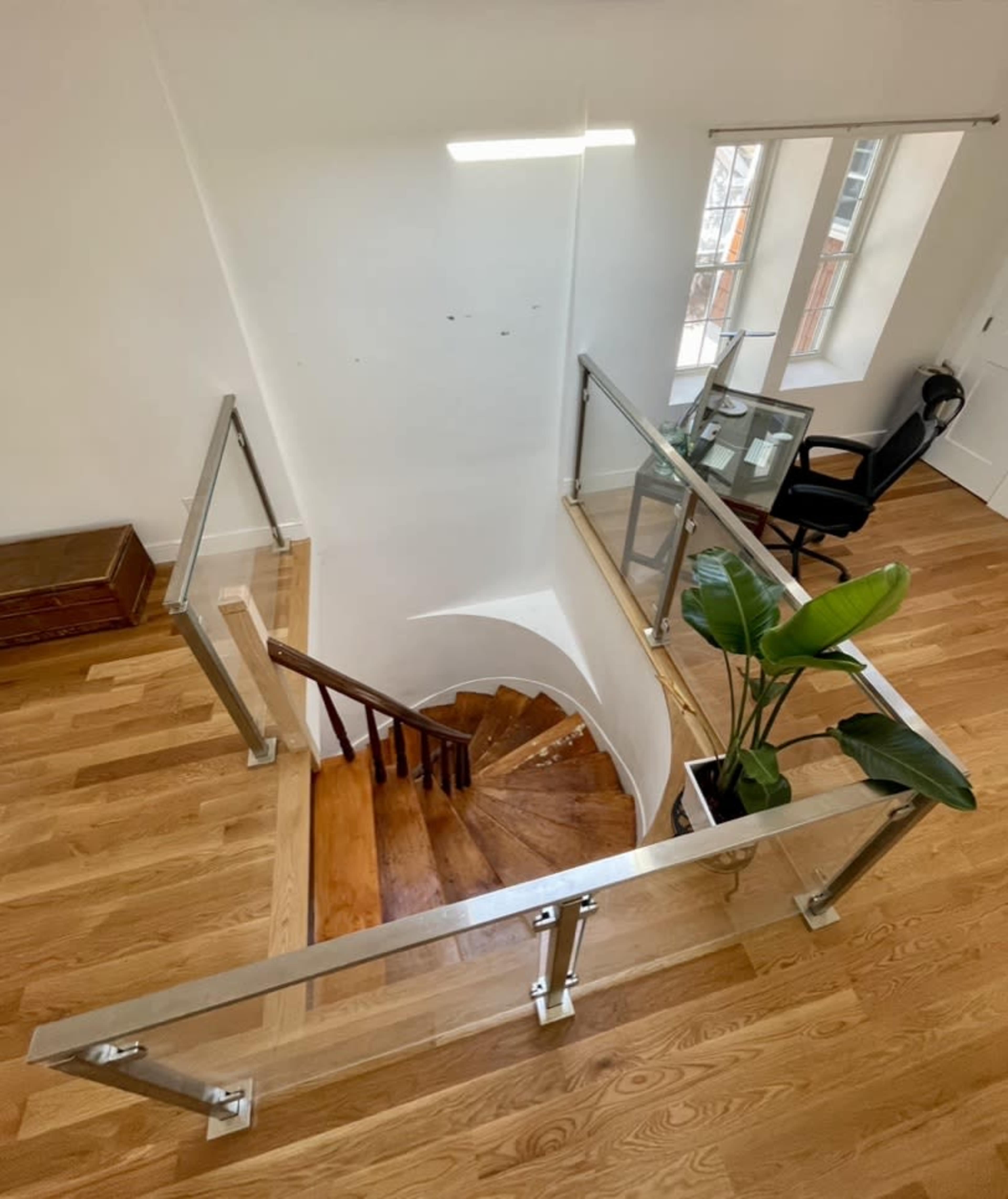 A spiral staircase leading down, surrounded by a glass railing, with a small desk and chair visible in the room at the bottom.