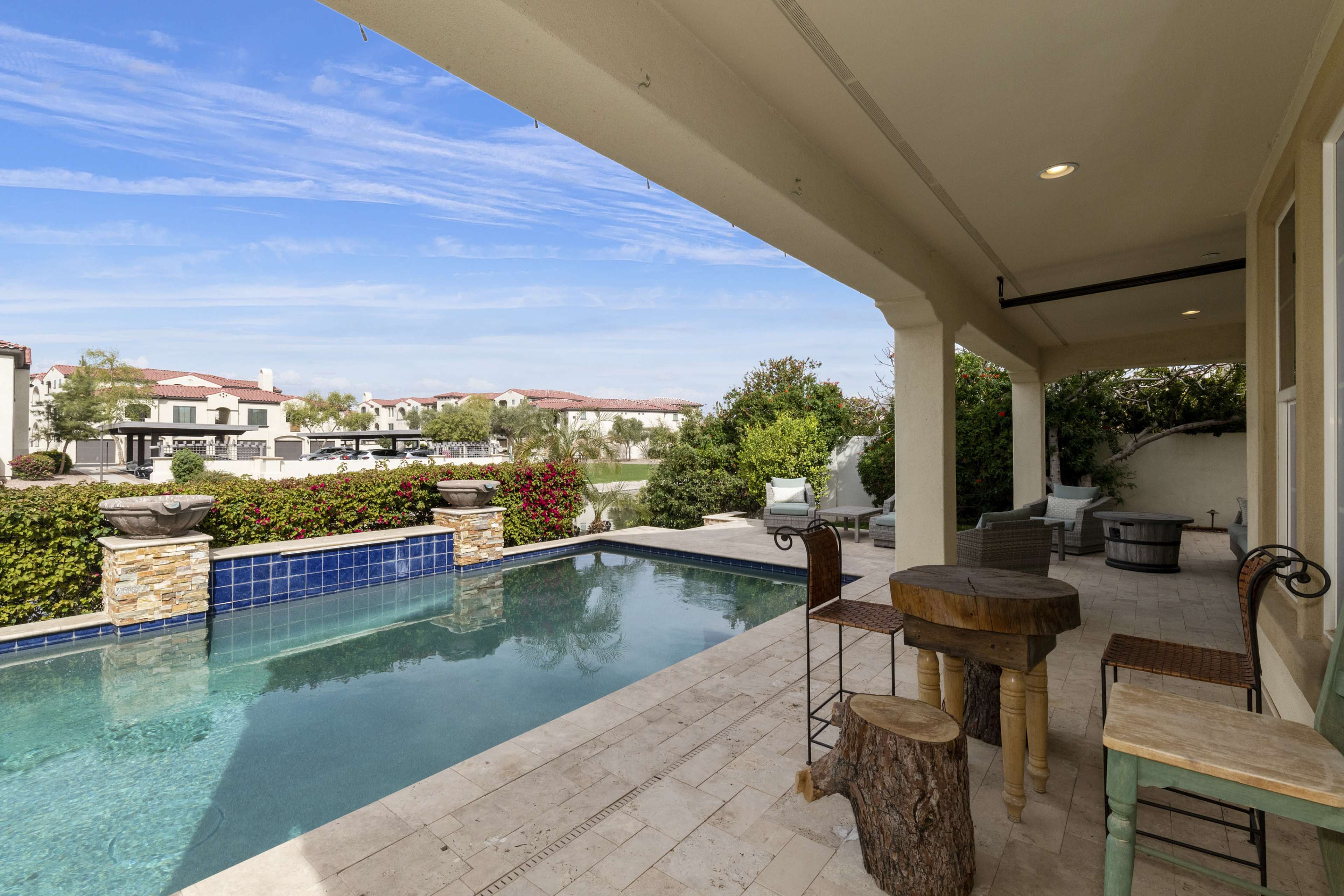 A swimming pool sits beside a covered patio adorned with wooden furniture, overlooking landscaped gardens and residential buildings in the background.