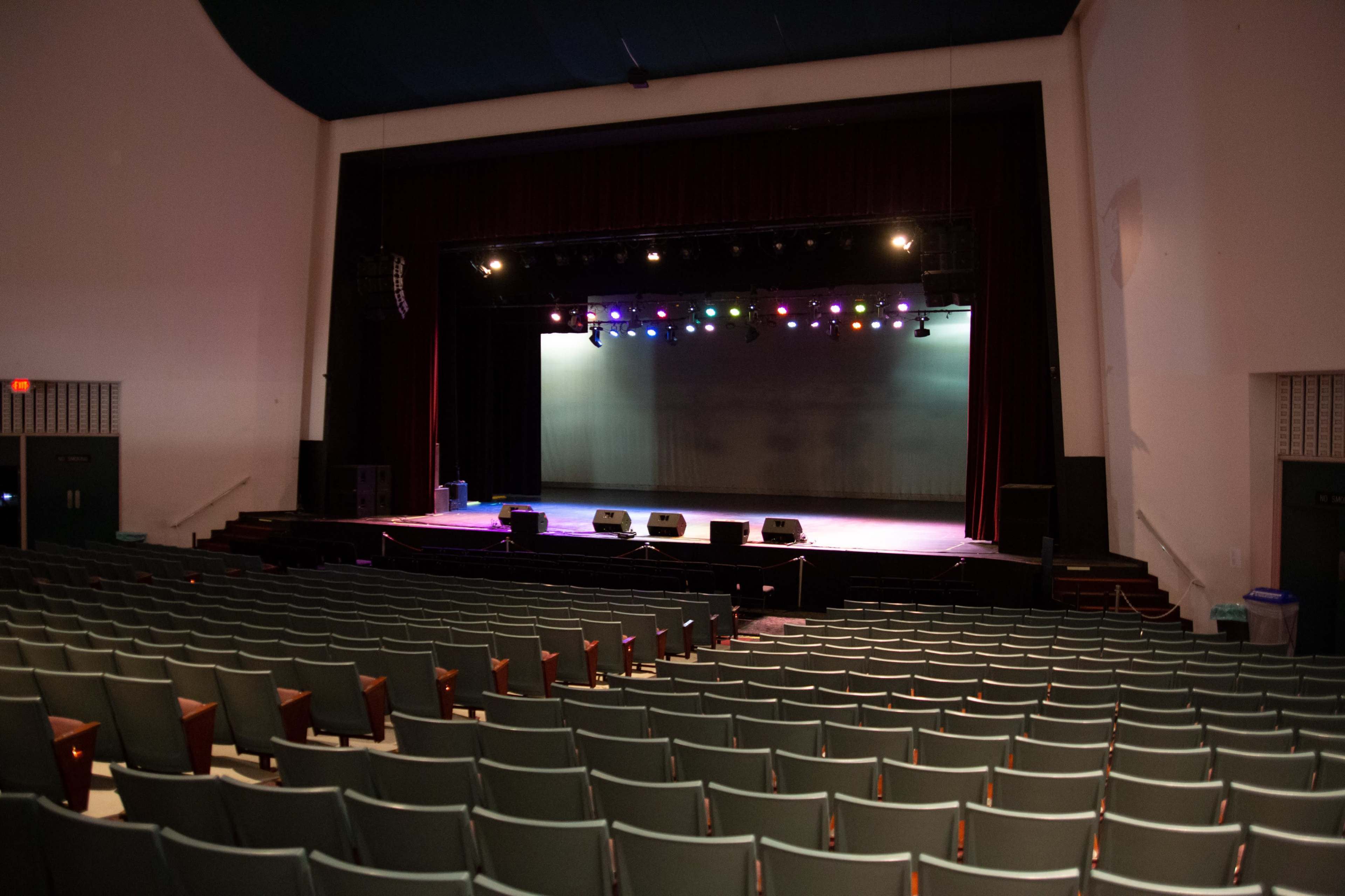 An empty theater with rows of green chairs facing a stage that has colorful lights and sound equipment set up.
