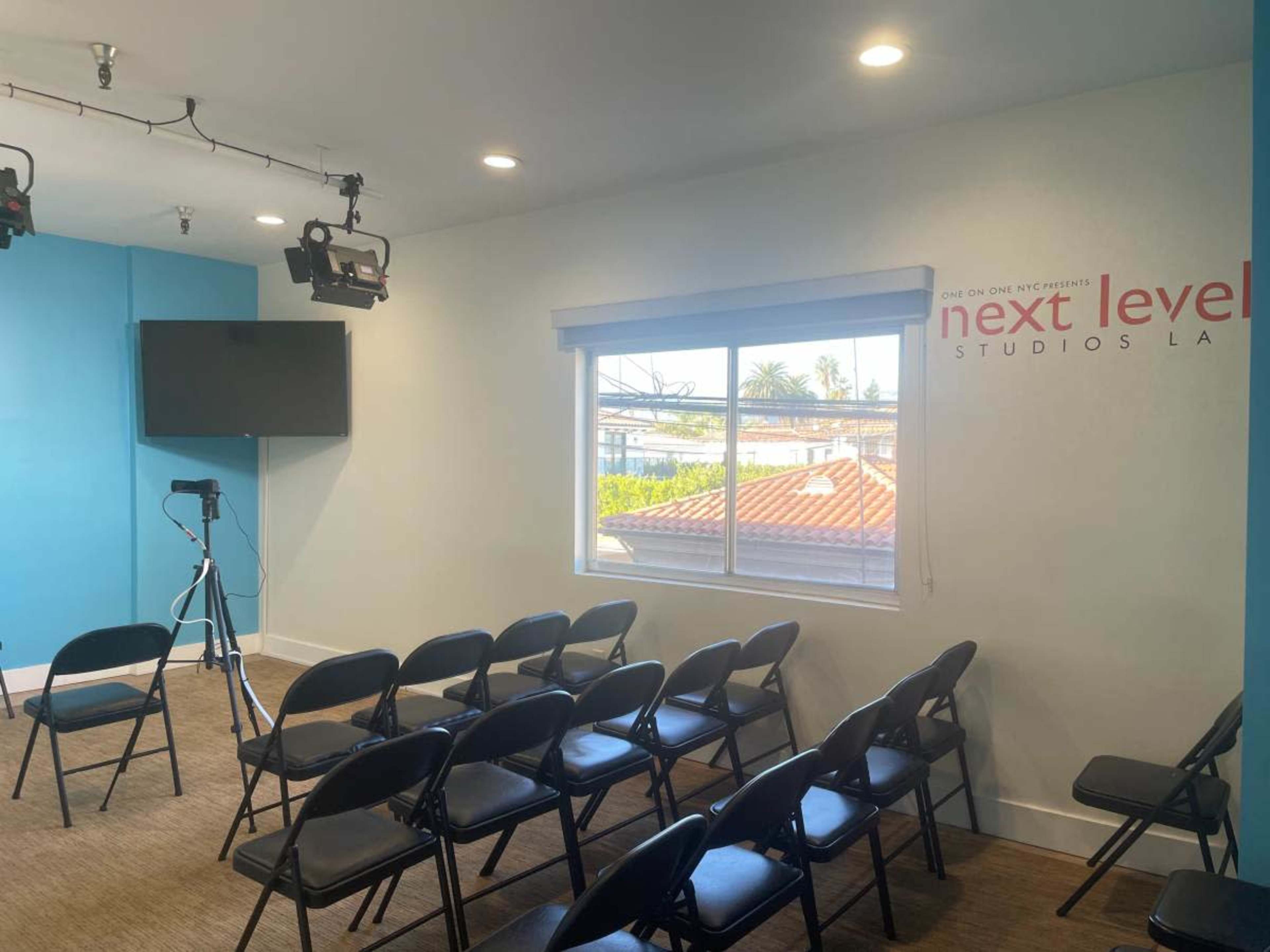 The image shows a small meeting room with rows of black chairs, a television mounted on the wall, and a sign that reads "next level studios LA."