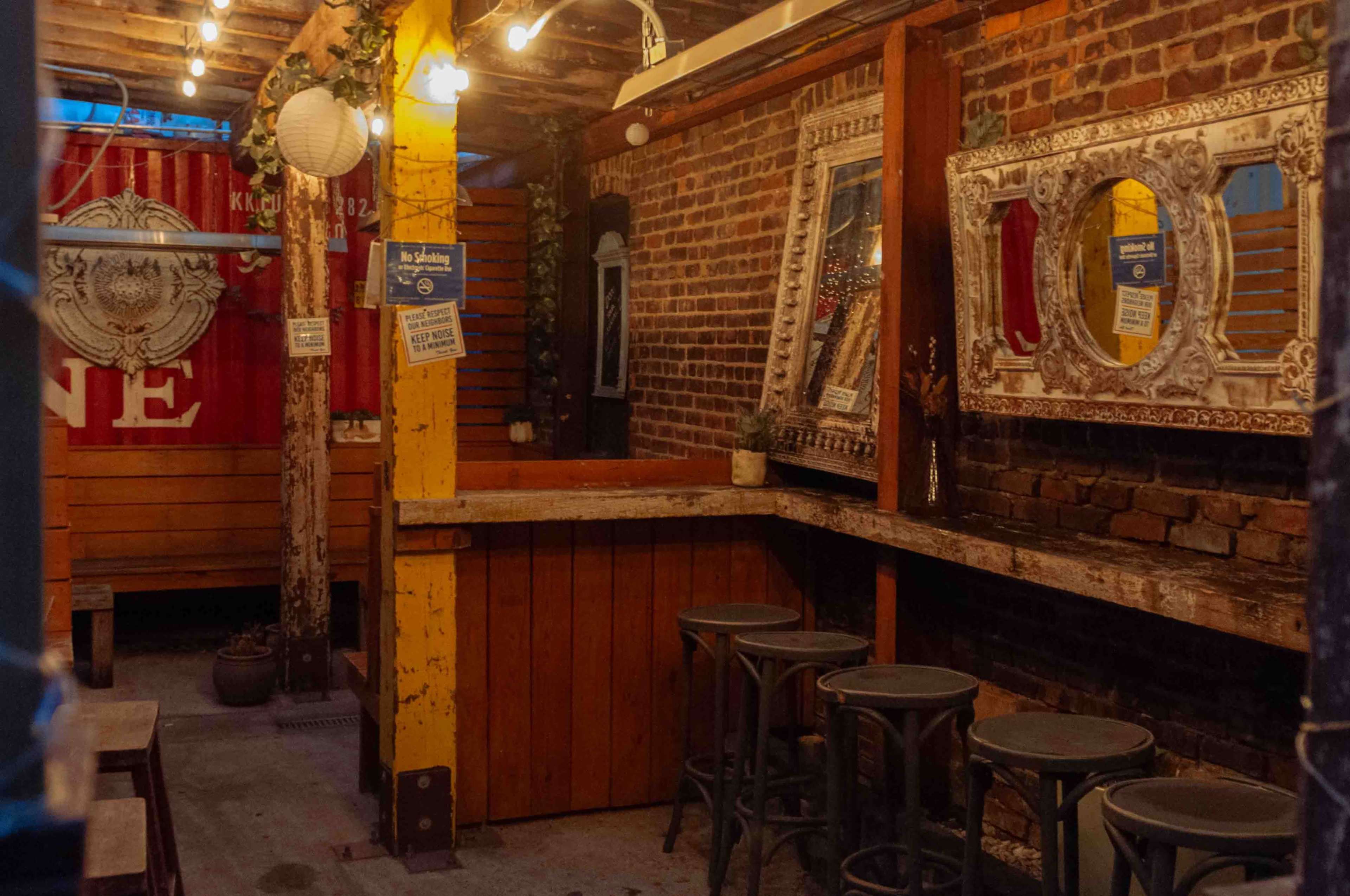 The image shows an intimate bar area with wooden benches, metal stools, and brick walls decorated with mirrors and string lights.
