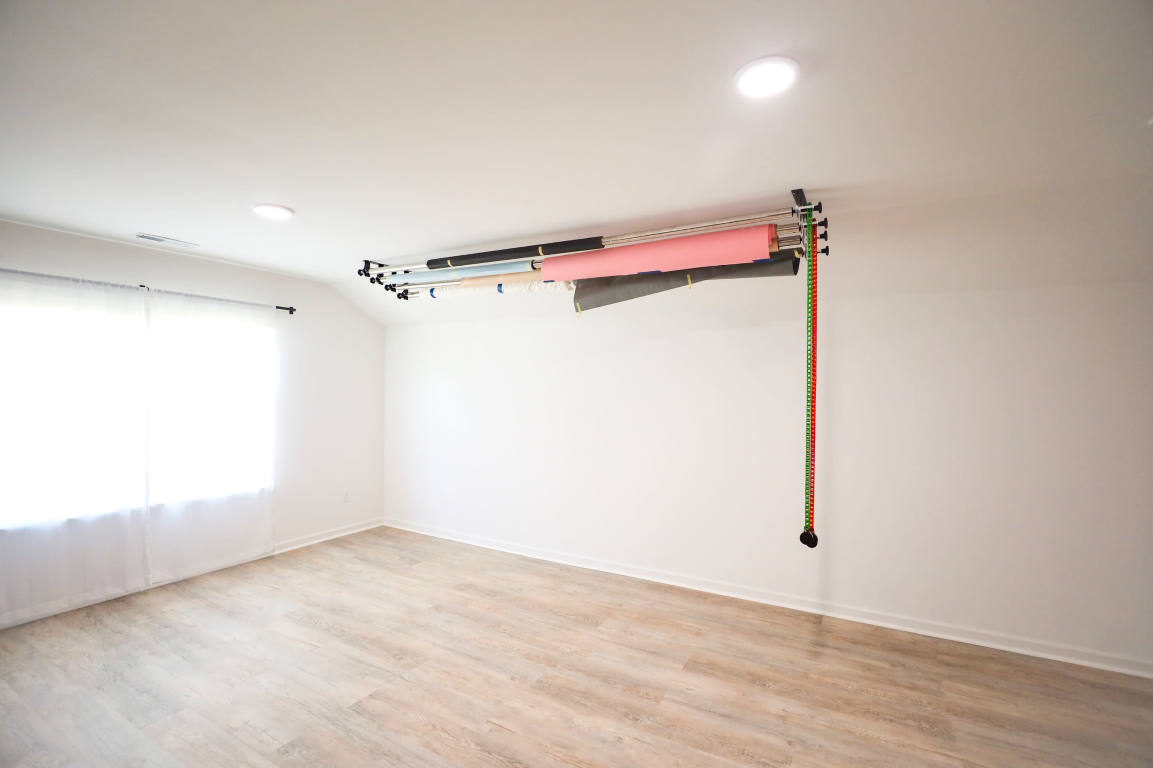 The image shows a spacious, empty room with wooden flooring and a window covered by a light curtain, featuring a wall-mounted rack holding colorful rolls of material at the ceiling.