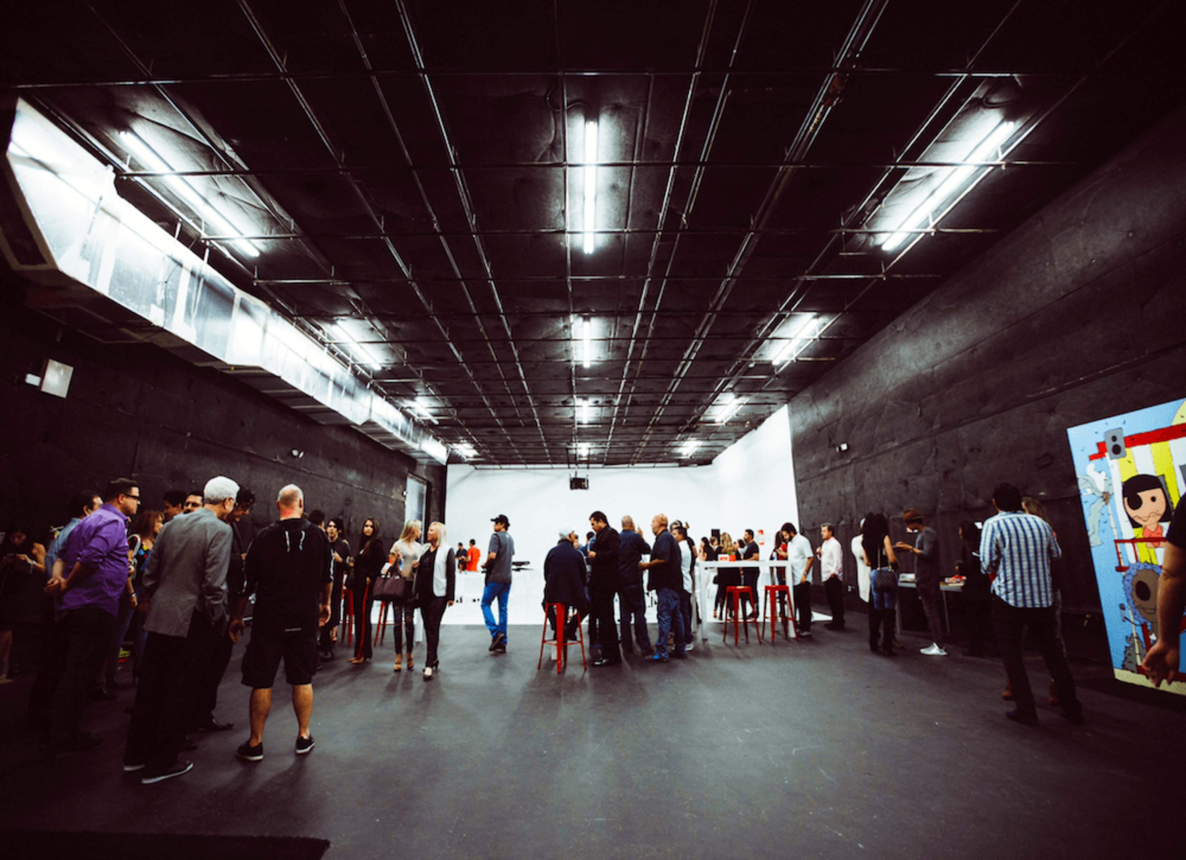 A group of people are mingling in a large, empty studio space with dark walls and a brightly colored mural.