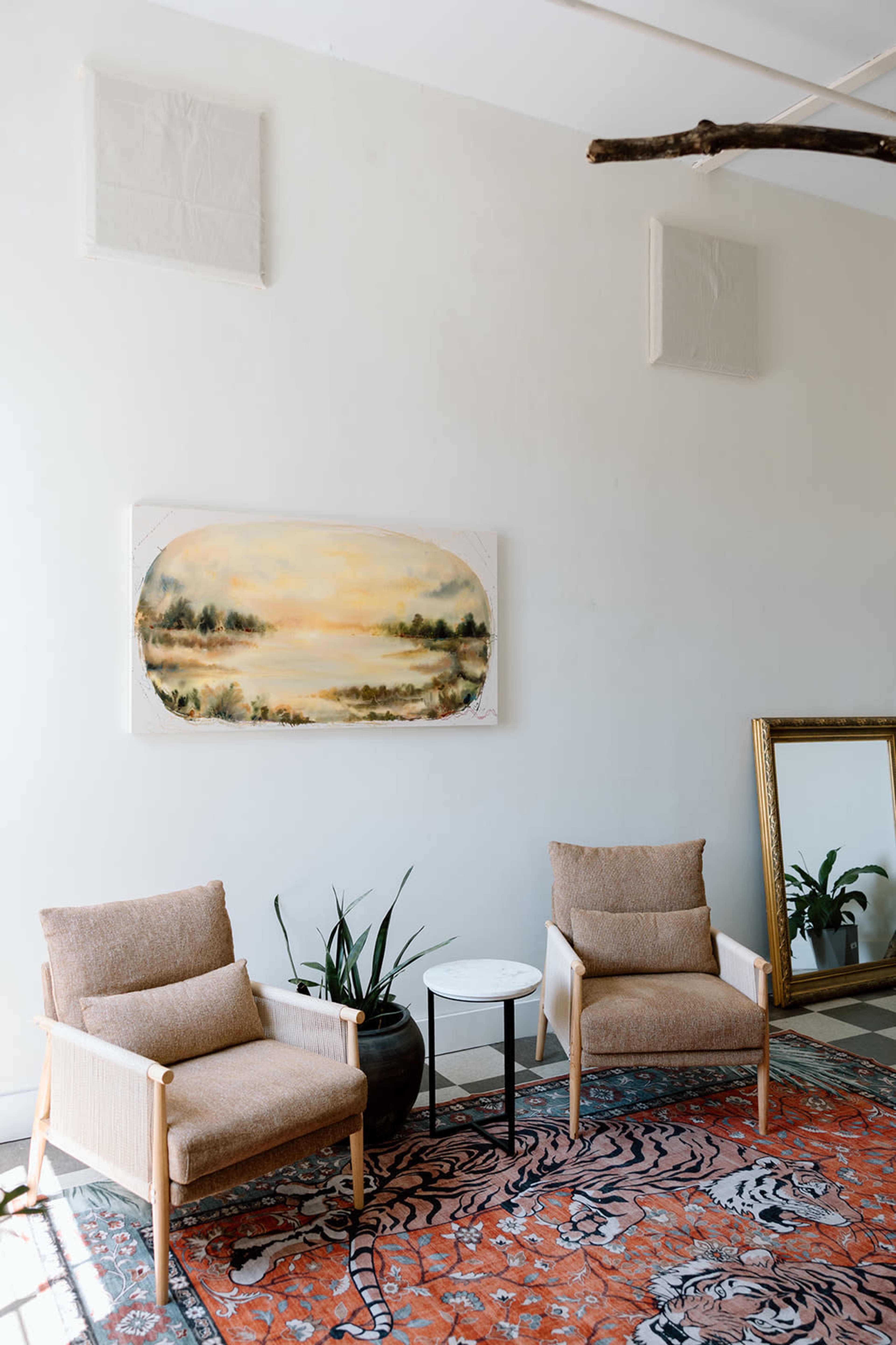 The scene features two armchairs flanking a small table on a patterned rug, with a large landscape painting and a mirror on the white wall.