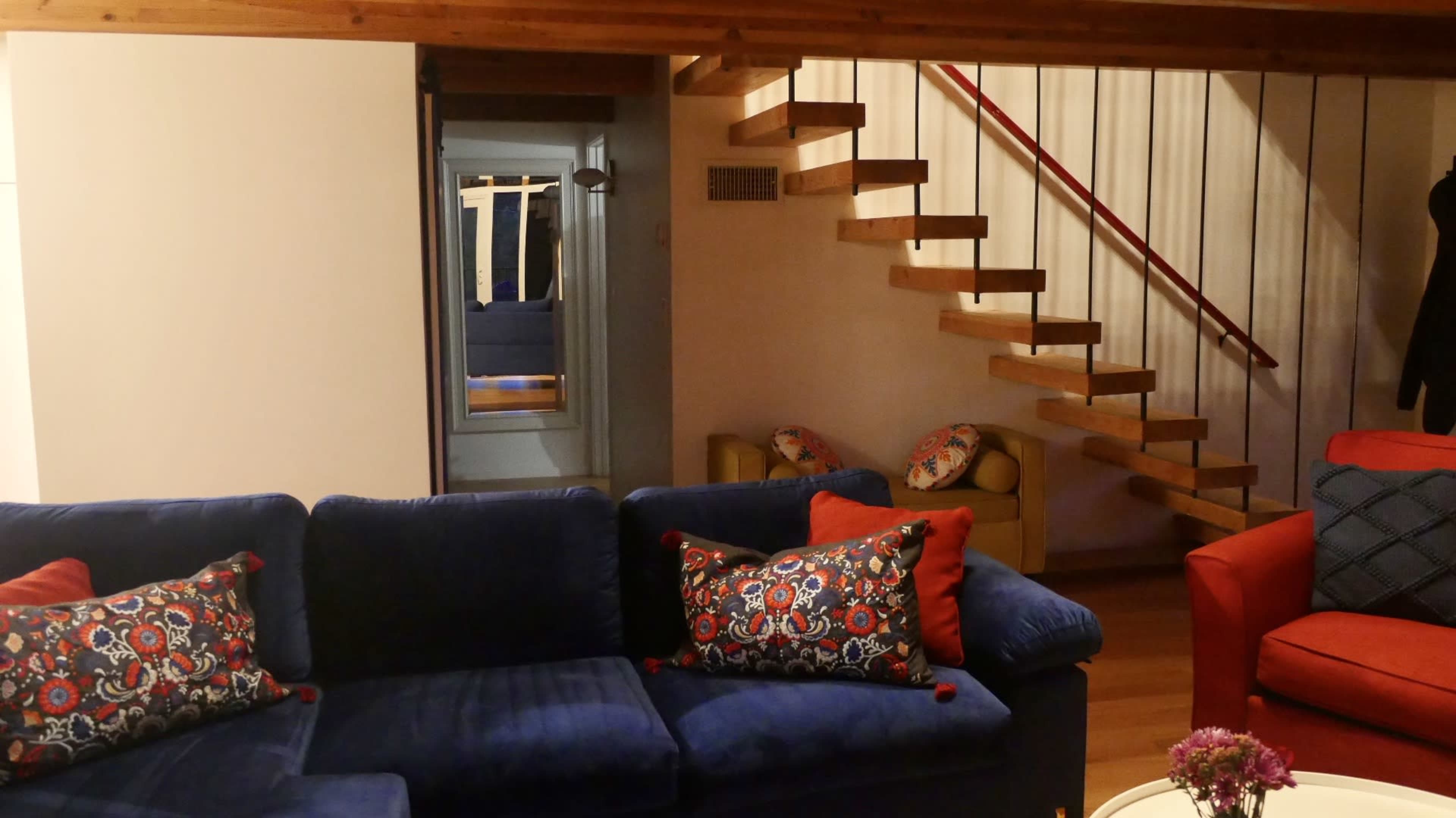 The image shows a cozy living area with a blue couch, red armchair, and a staircase leading to an upper level.