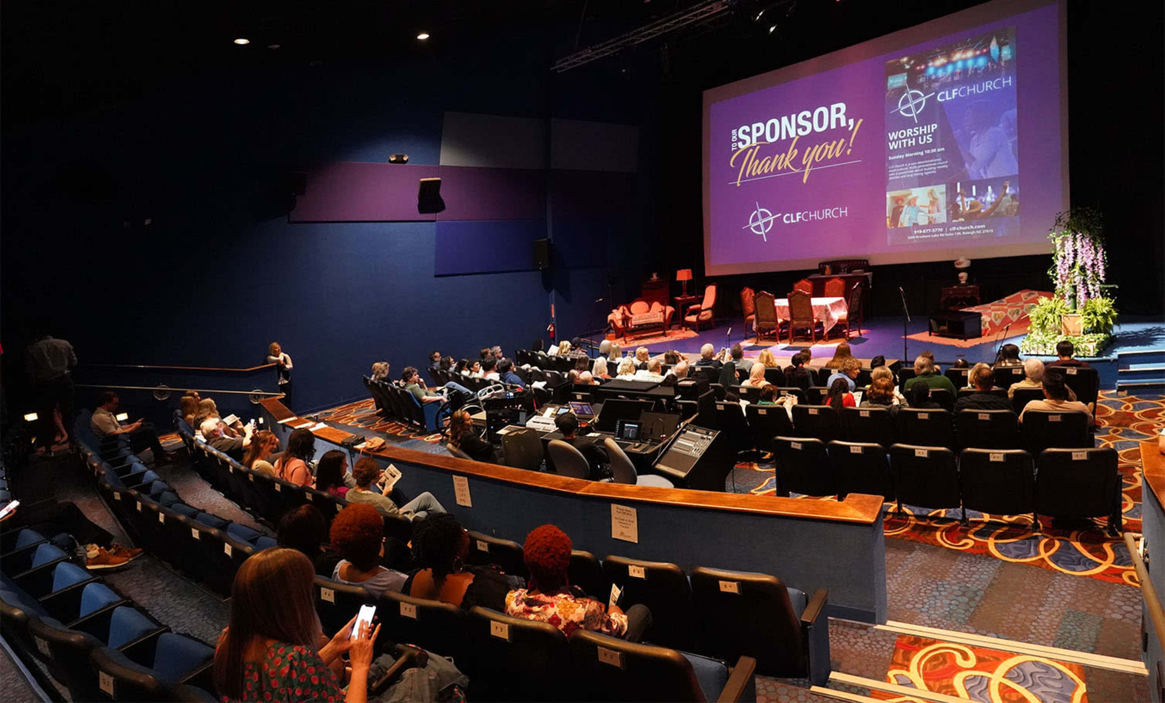 An audience is seated in a theater, facing a stage with a large screen displaying a message of thanks to sponsors for an event.