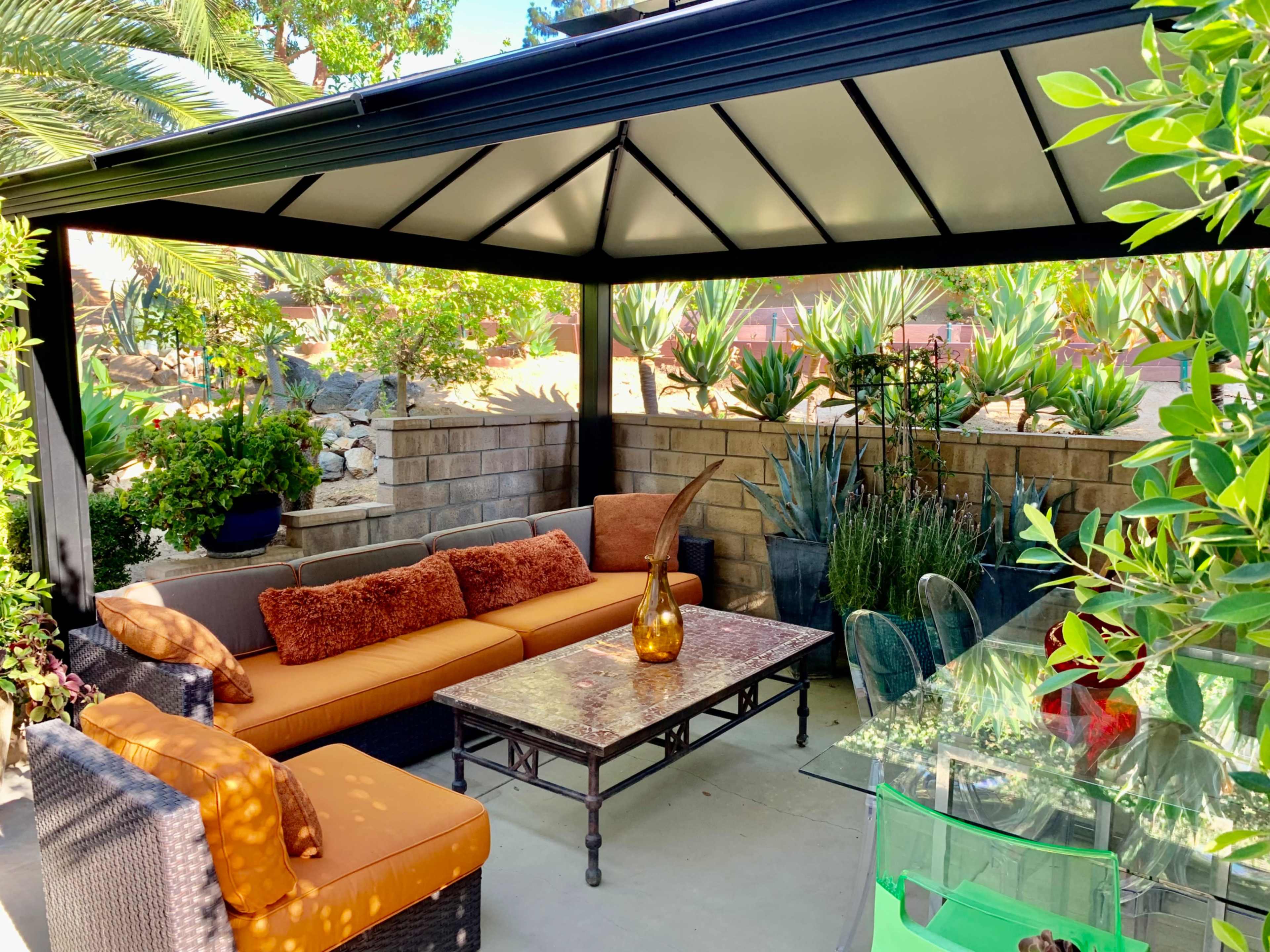 A shaded outdoor seating area with a sofa, a glass table, and various potted plants around a stone wall.