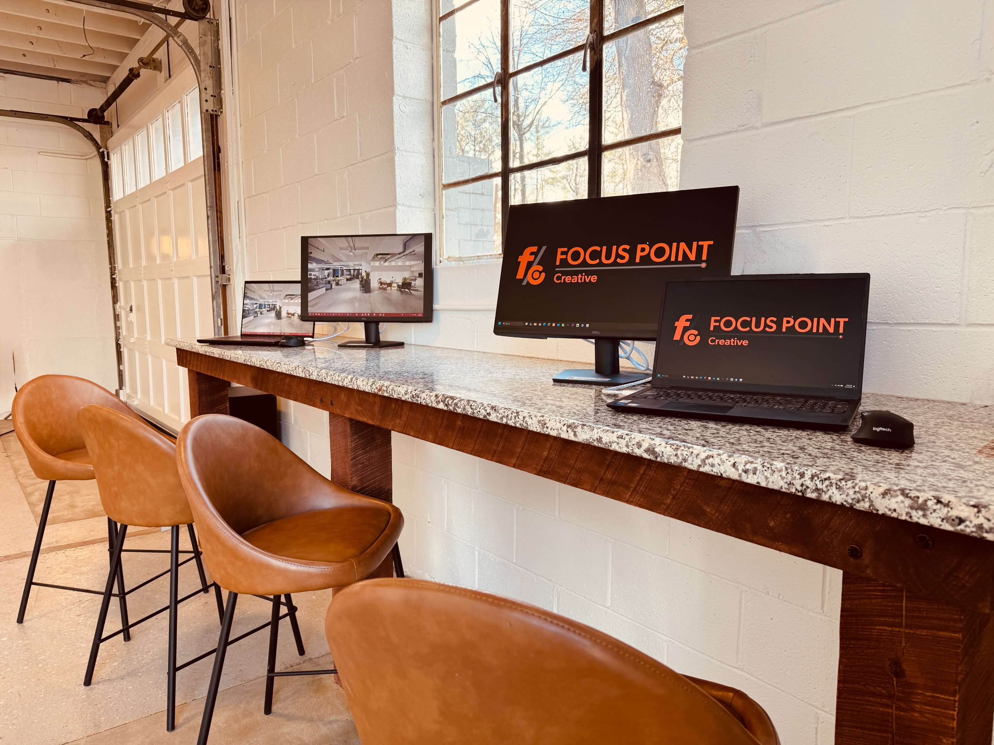 The image shows a workspace with two computer monitors and a laptop on a wooden counter, alongside three brown barstools.
