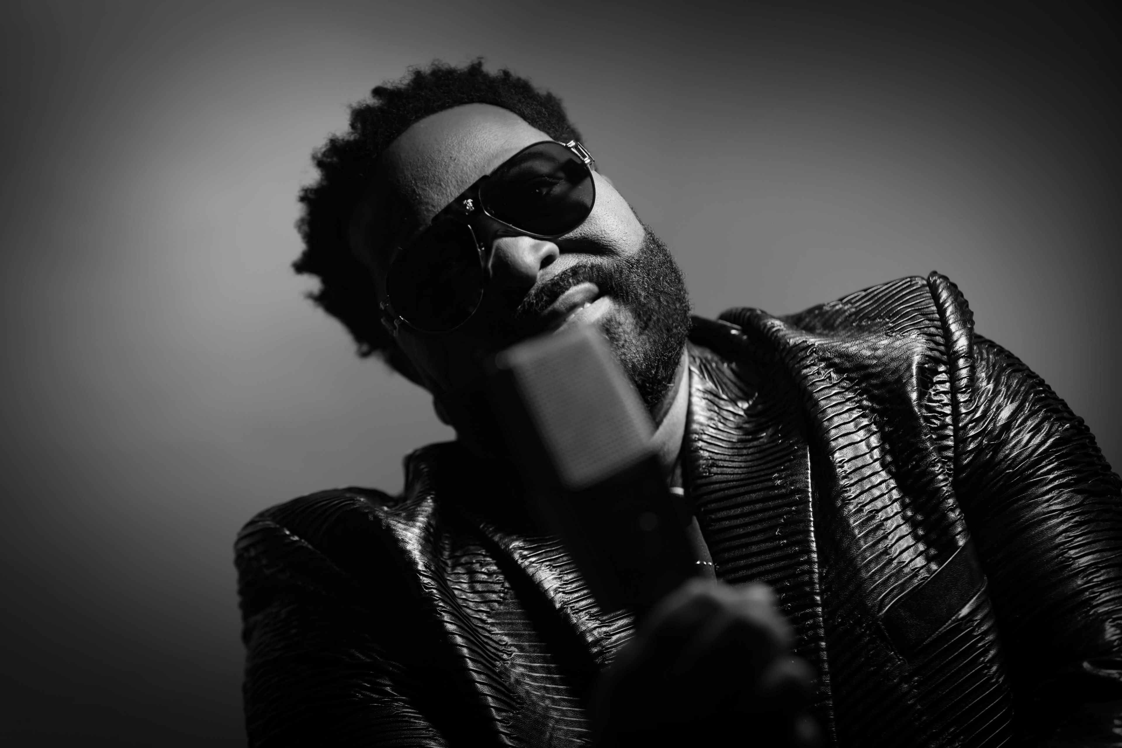 A man in sunglasses holds a retro microphone while wearing a shiny, textured jacket against a dark background.