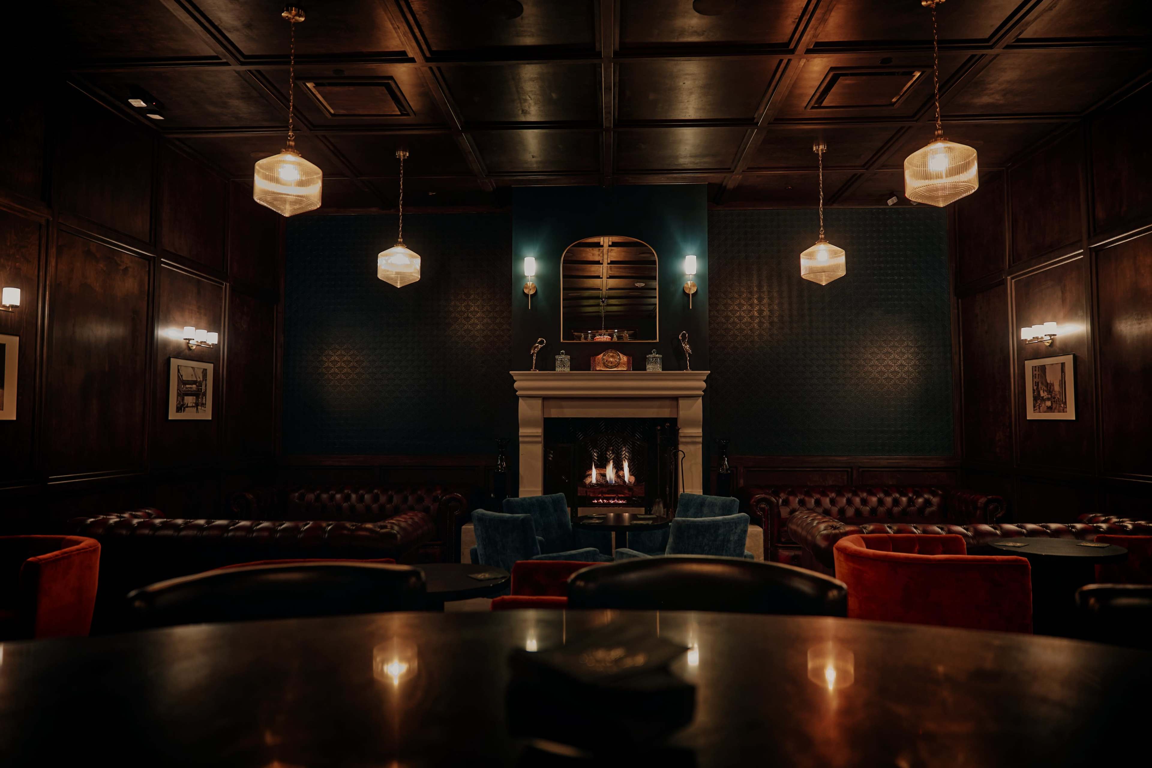 A dimly lit lounge with leather sofas and a central fireplace, complemented by hanging pendant lights.