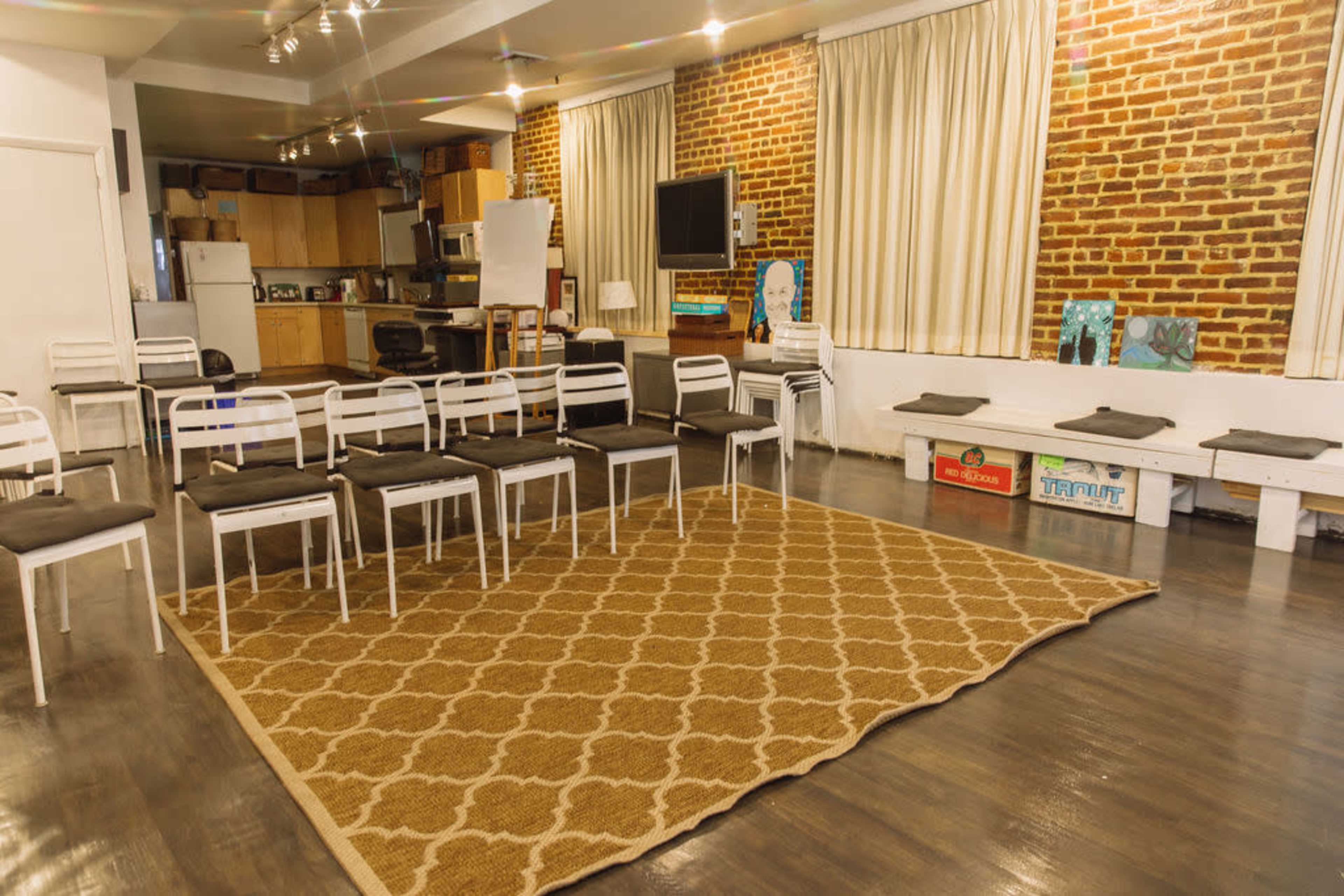 The image shows a room with a circular arrangement of white chairs on a patterned carpet, with brick walls and large windows draped with curtains.