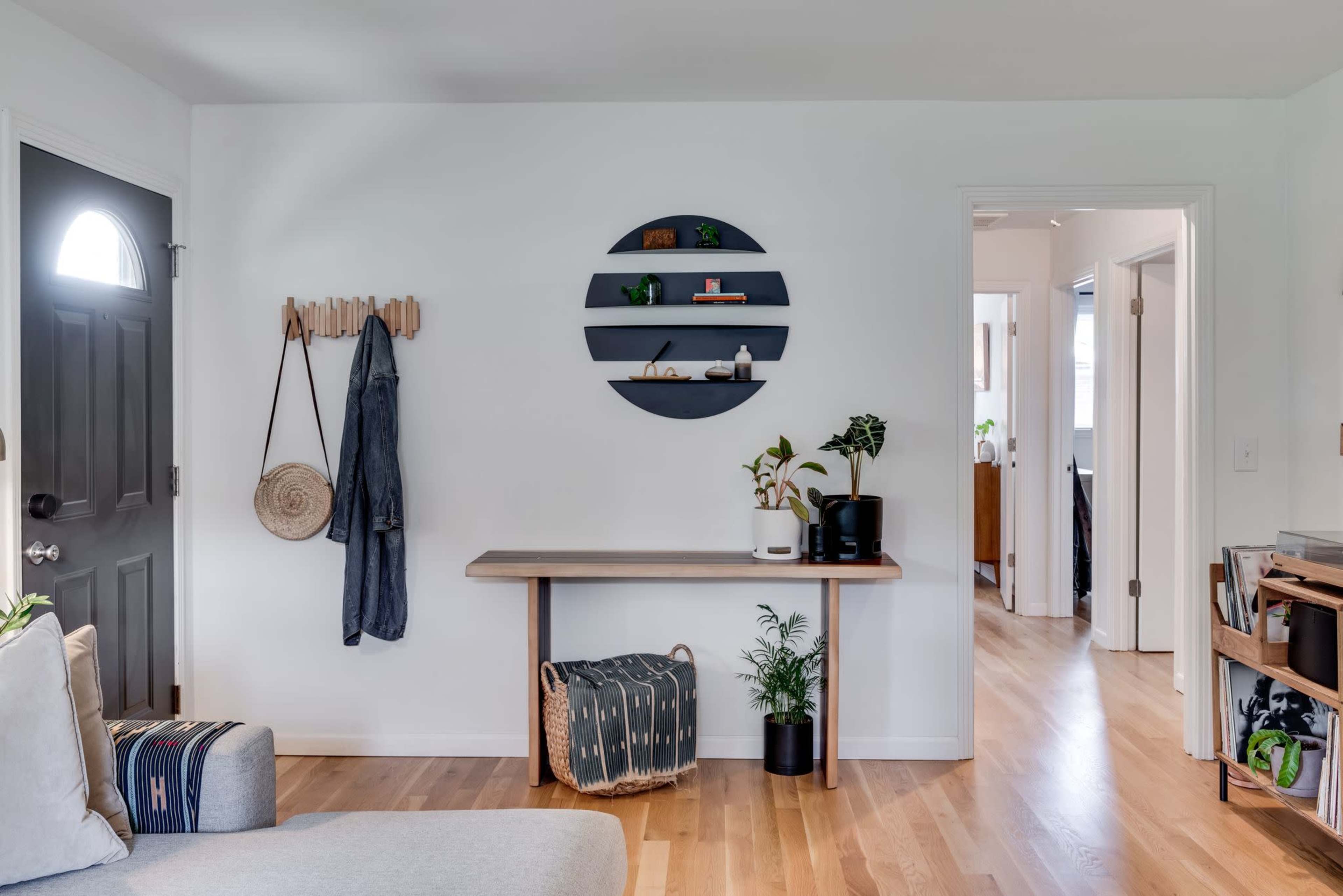A modern living space with a small wall-mounted shelf, a coat rack, and a wood table against a white wall, featuring a plant and decorative items.