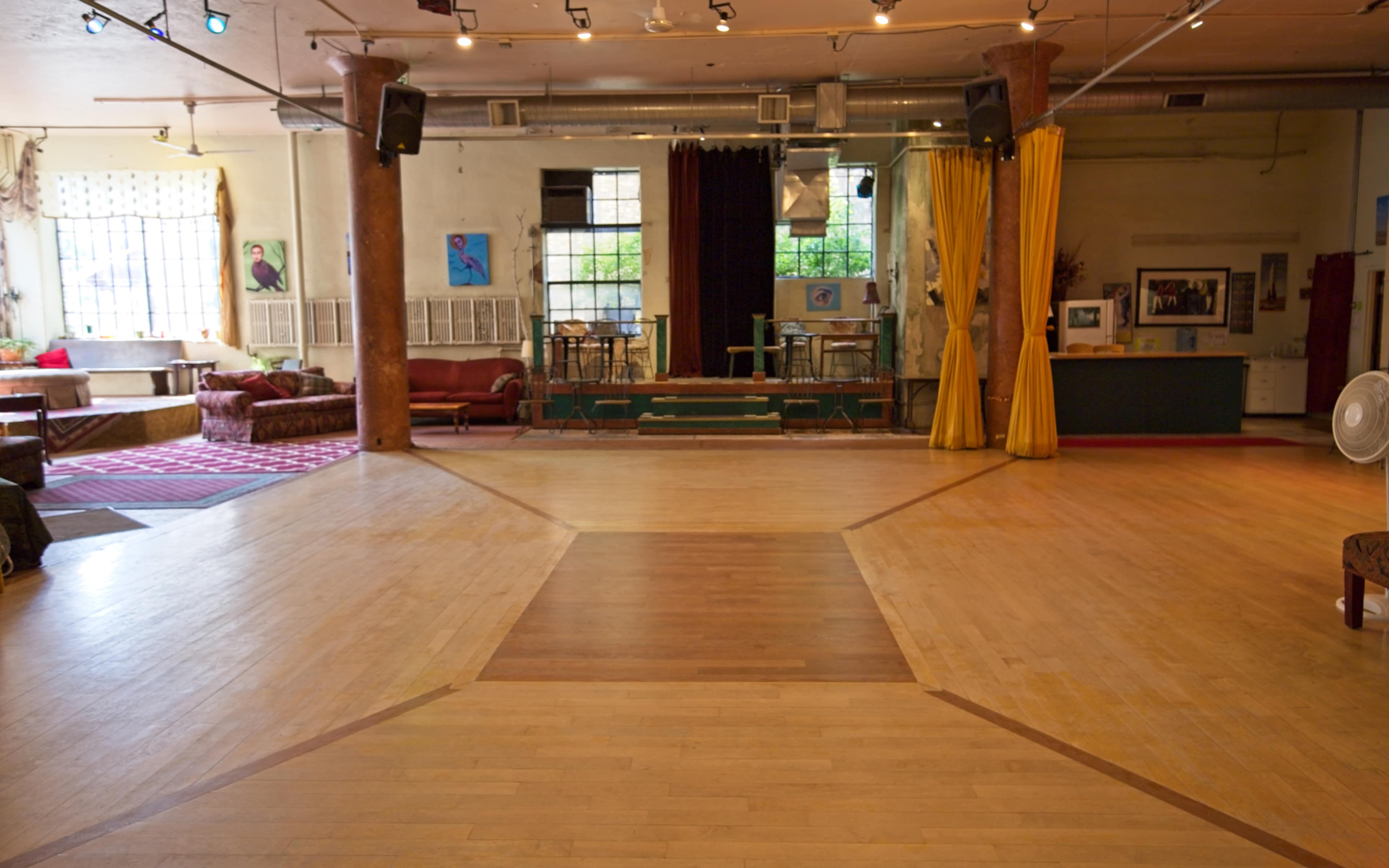 An empty dance studio featuring a wooden floor, seating areas, and a small stage at the far end.