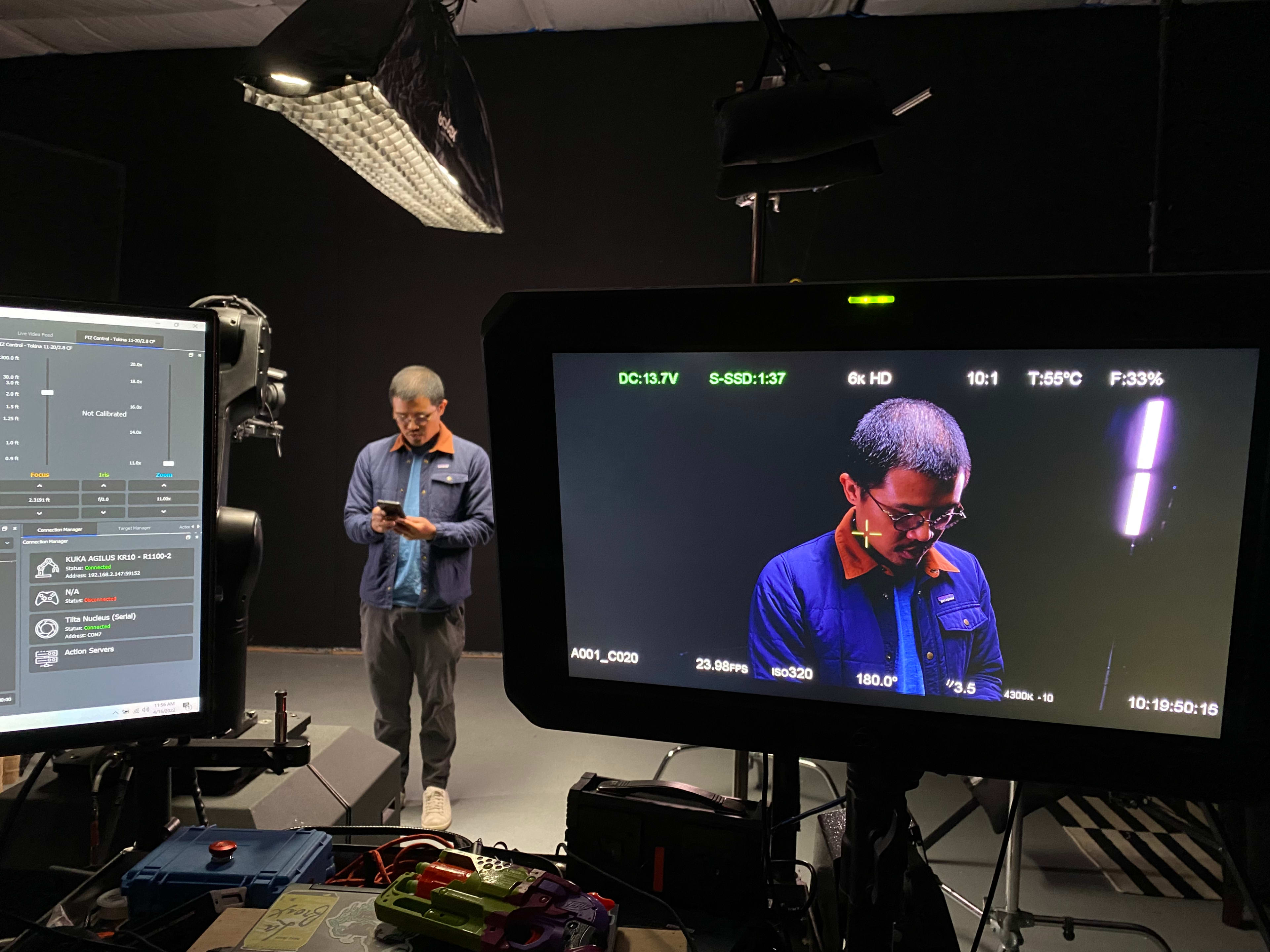 A man stands in a studio, focused on his smartphone, while a camera feed displays his image on a monitor nearby.
