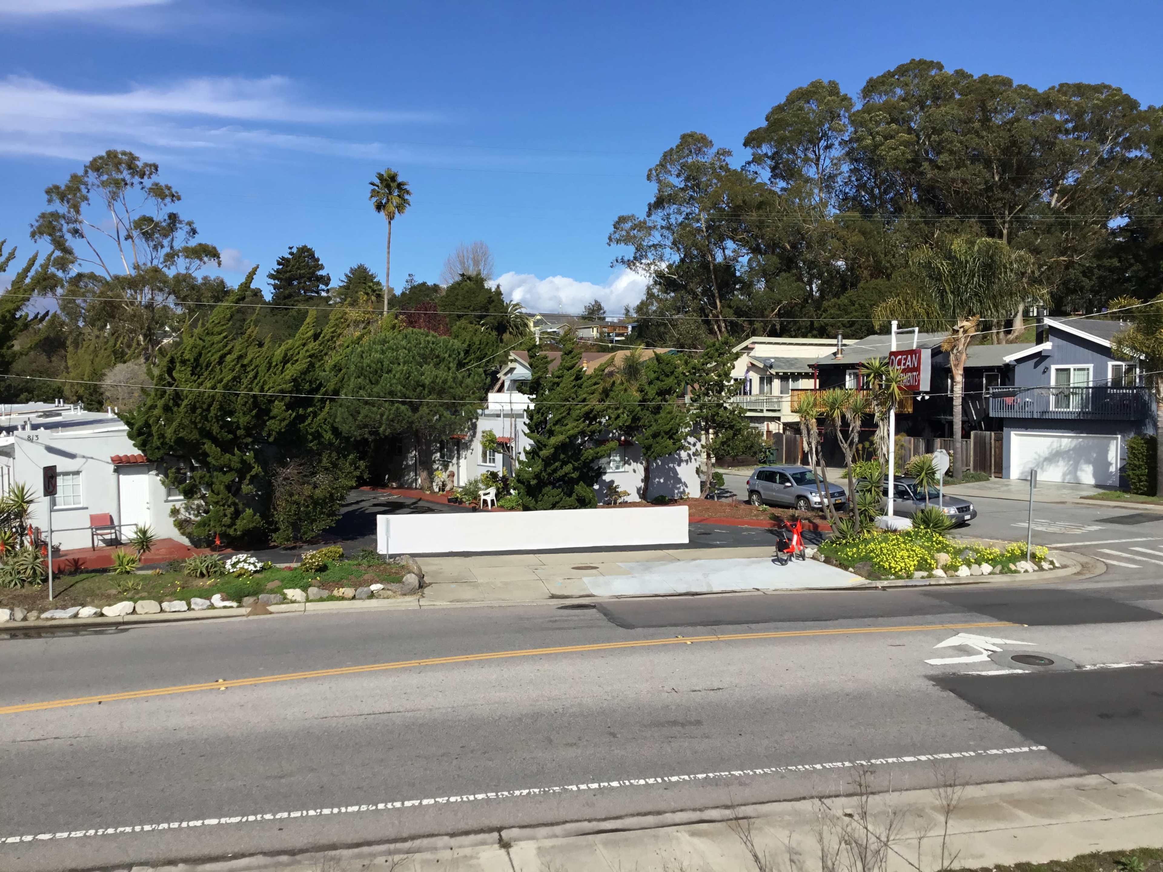 The image shows a street view of a neighborhood with residential buildings, palm trees, and landscaped areas beside a road.