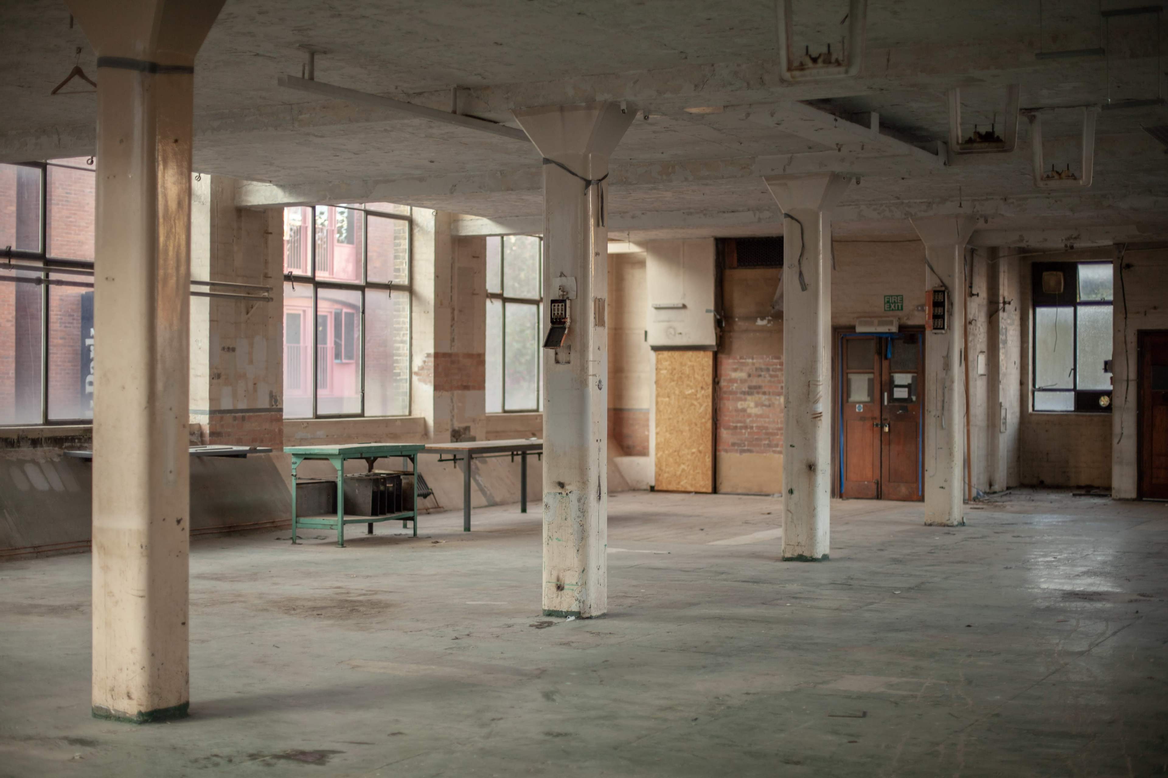 The image shows an empty industrial space with concrete pillars, large windows, and a few pieces of furniture against the walls.