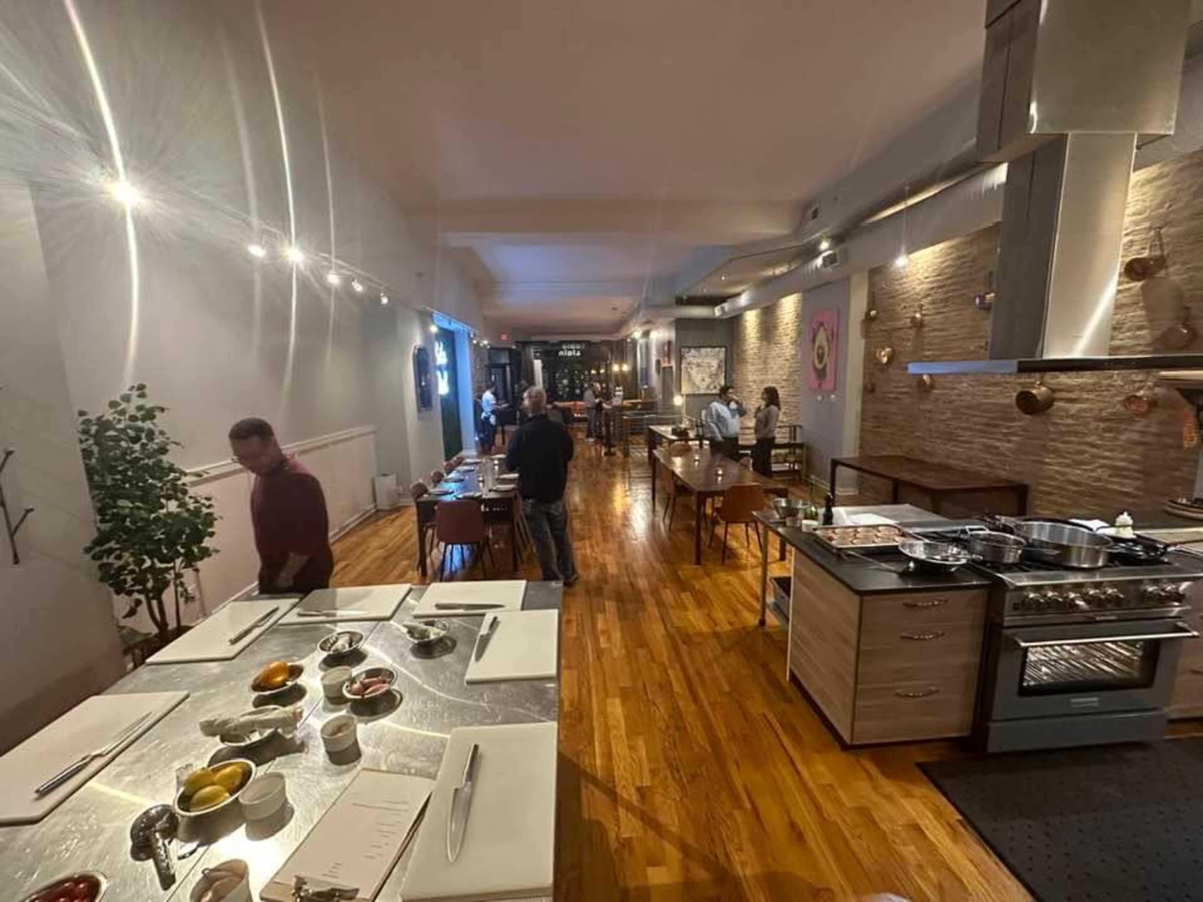A spacious restaurant interior features wooden floors, tables set for dining, and a cooking area with various ingredients and appliances.