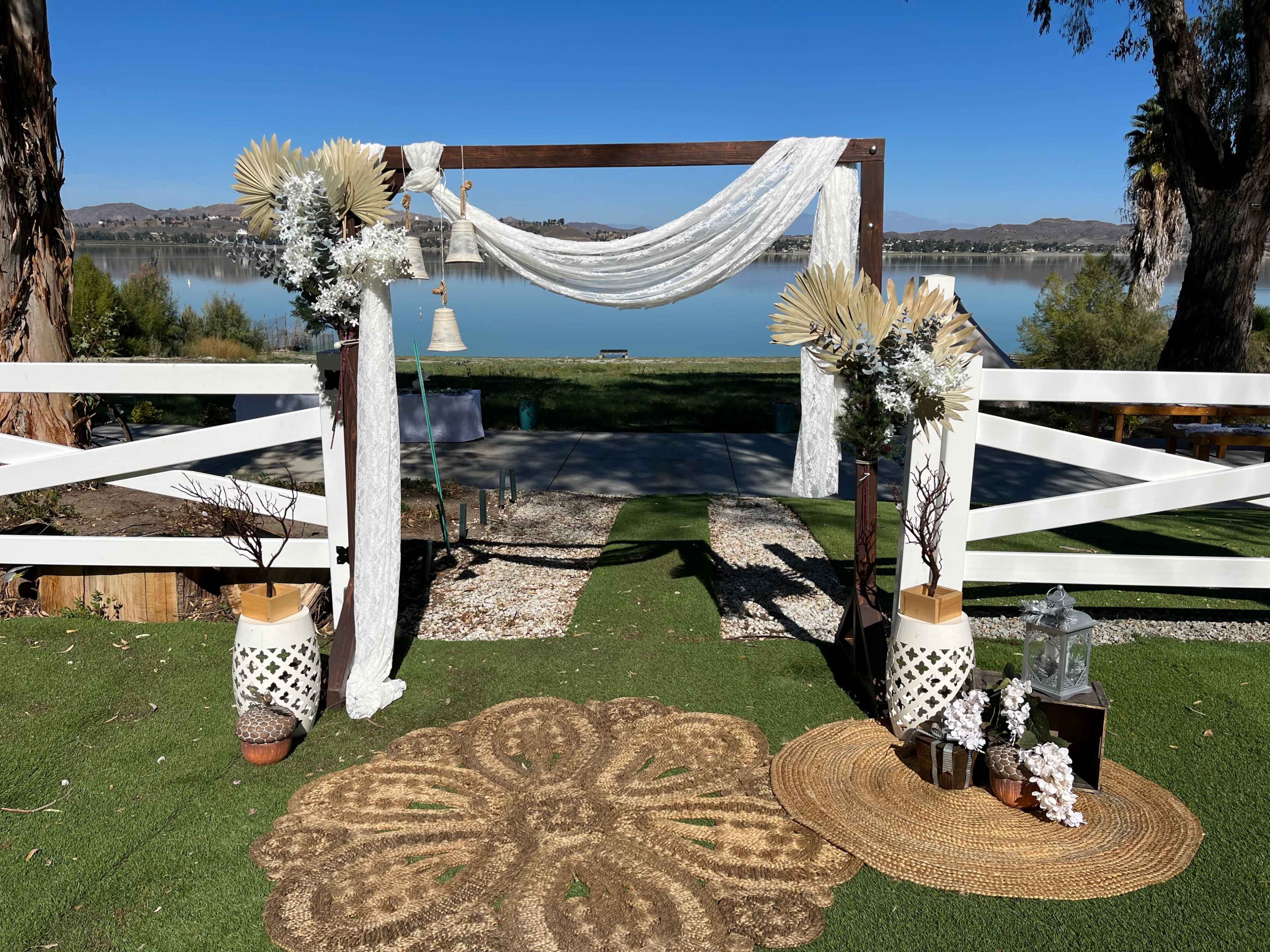 A decorative wedding arch set up by a lake, adorned with fabric drapes, dried foliage, and floral arrangements.