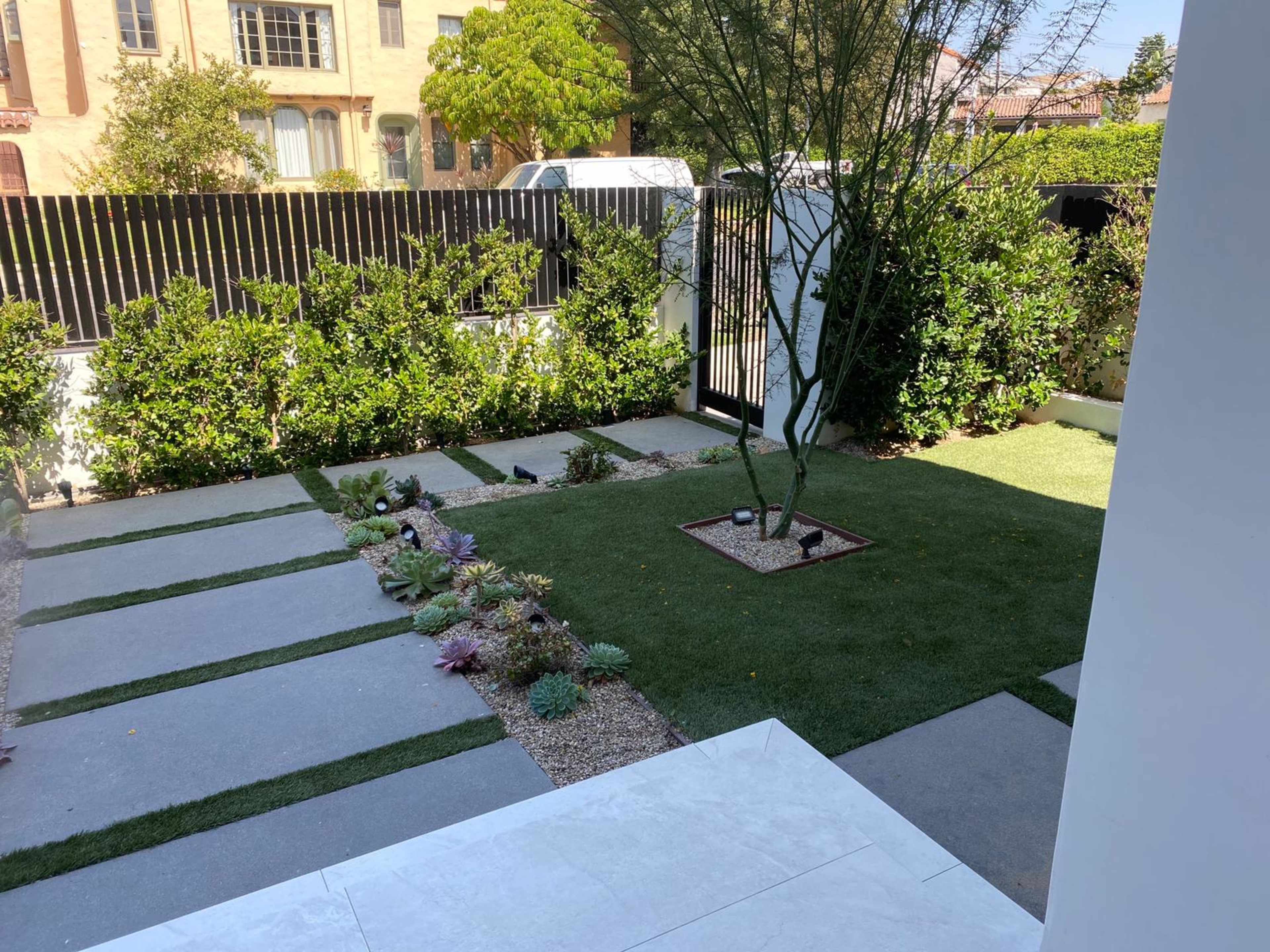The image shows a landscaped garden with stone pathways, grass areas, and various plants enclosed by a black fence.