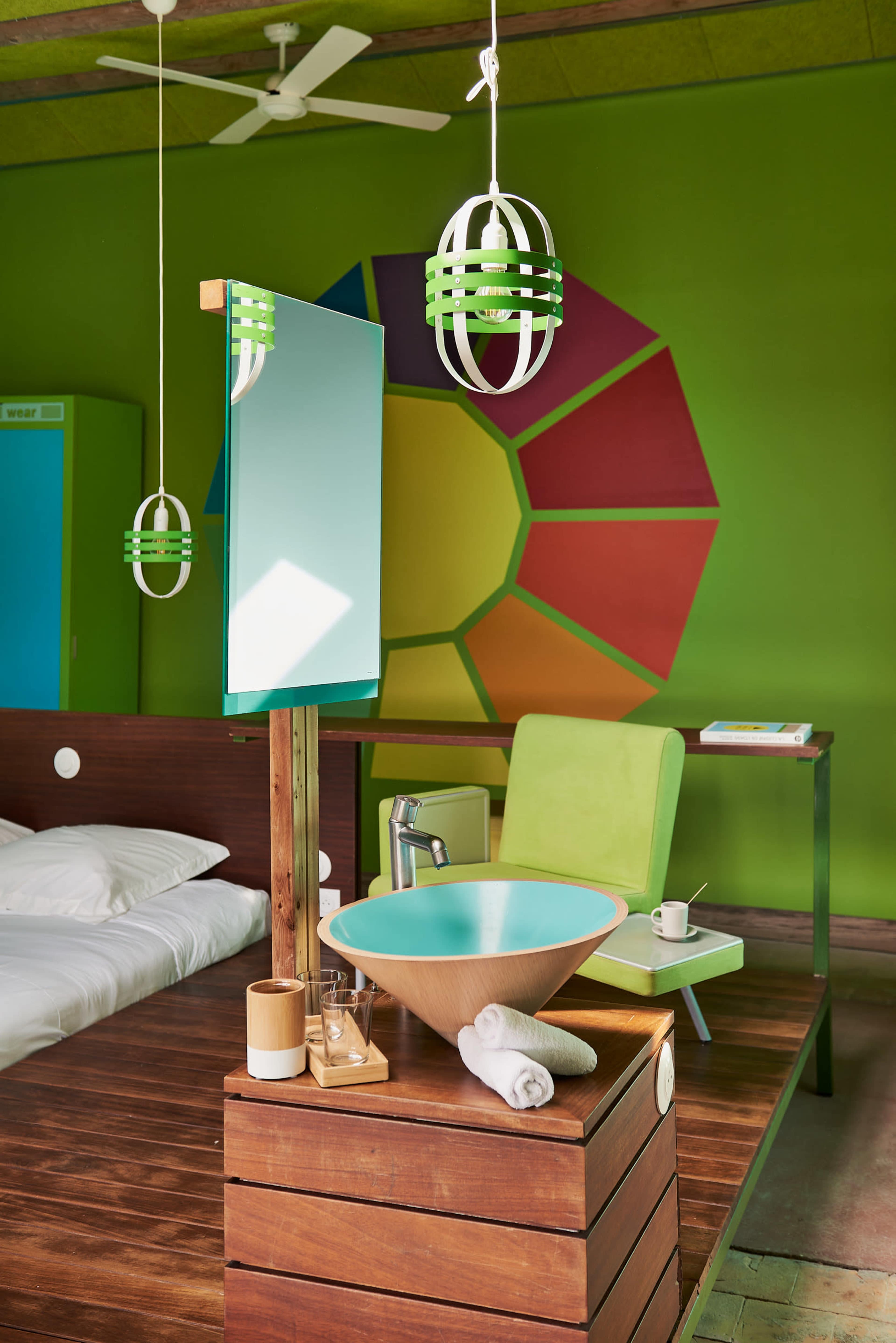 The image shows a colorful hotel room with a wooden bed, a green accent chair, a modern bowl sink on a wooden stand, and vibrant wall art in the background.