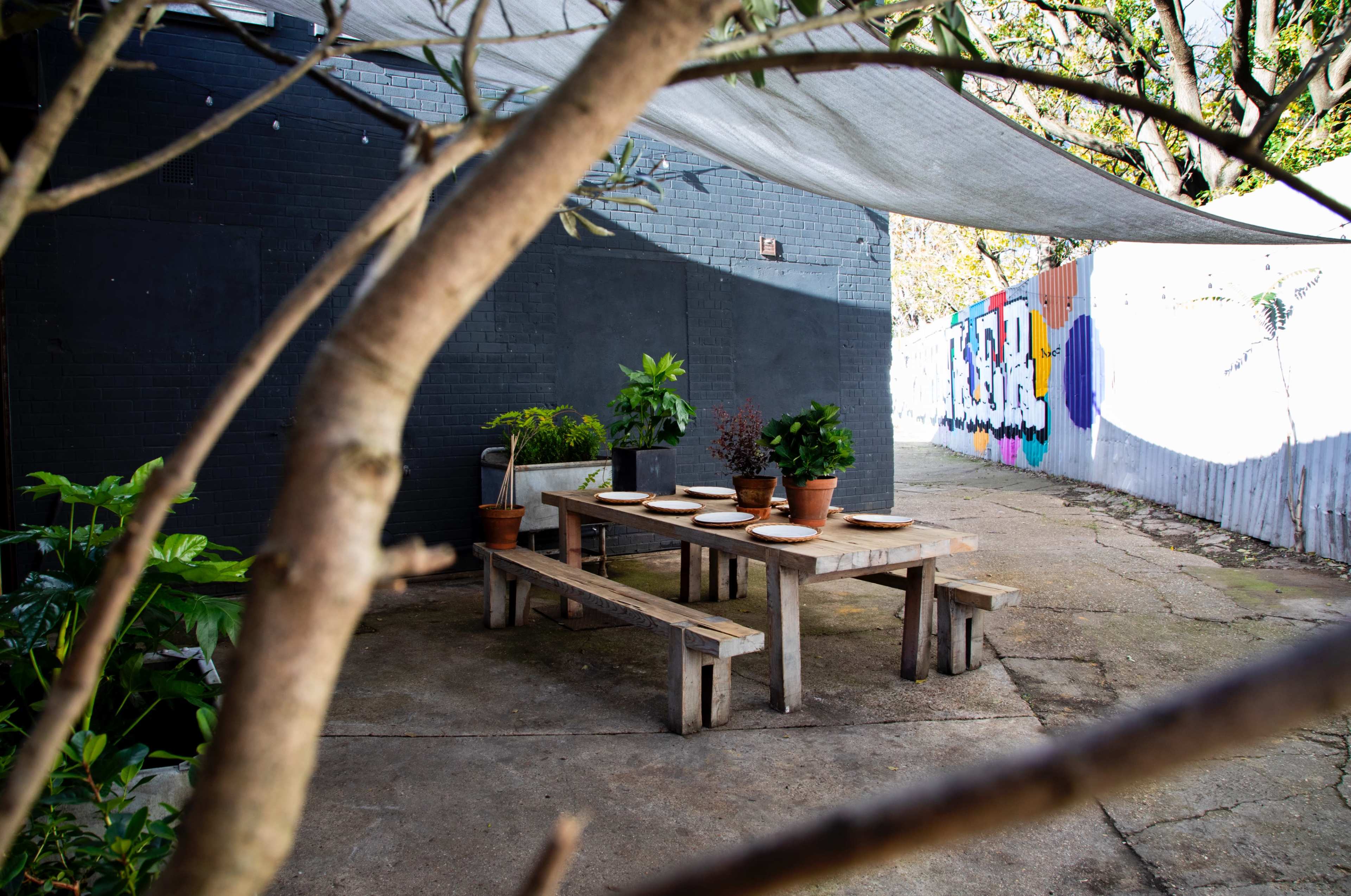 A wooden table with plates is set outside under a shaded area, surrounded by potted plants and a colorful mural in the background.