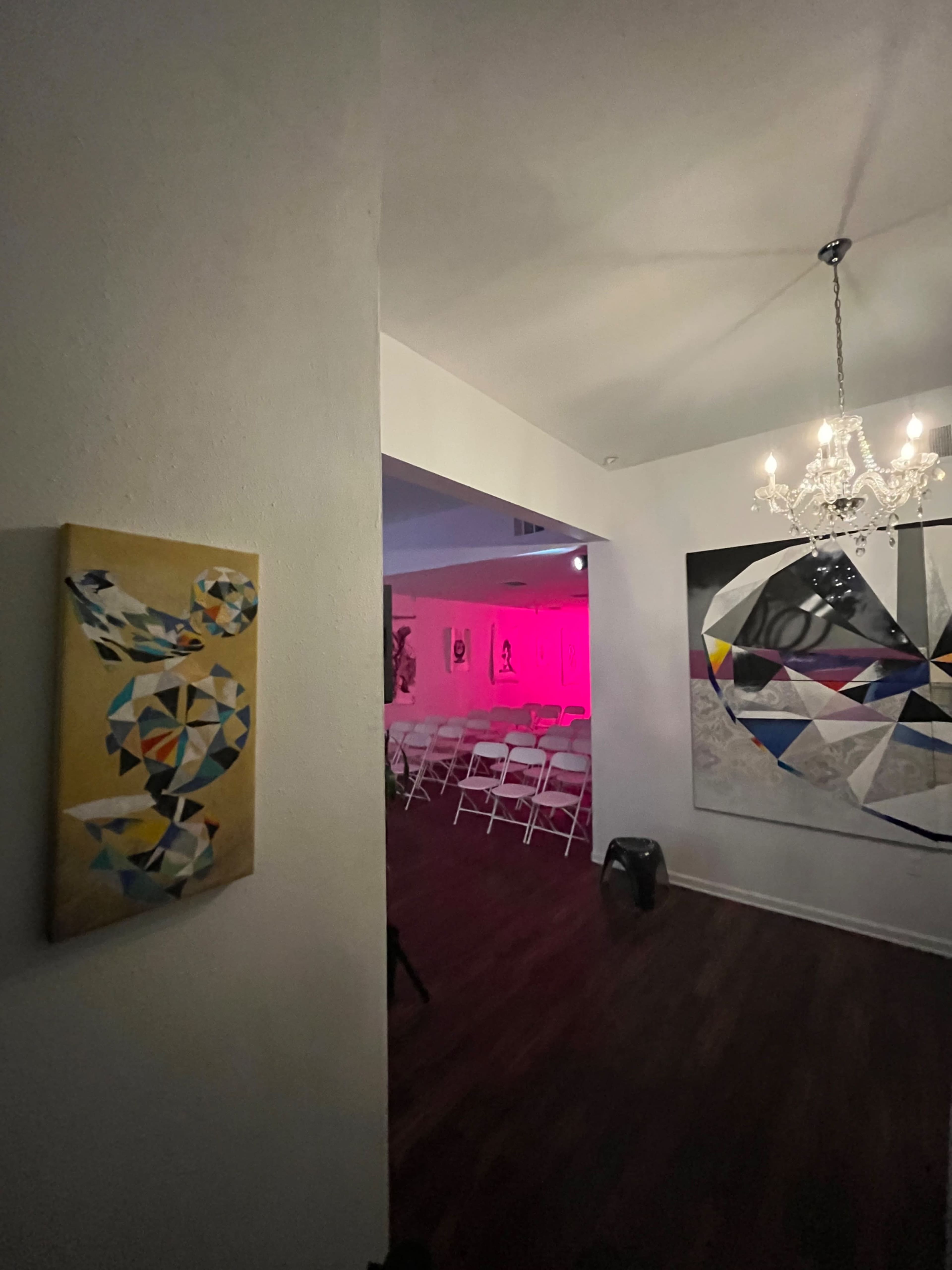 The image shows an art gallery with a geometric painting on the wall to the left and rows of white chairs set up for an event in the illuminated room ahead.