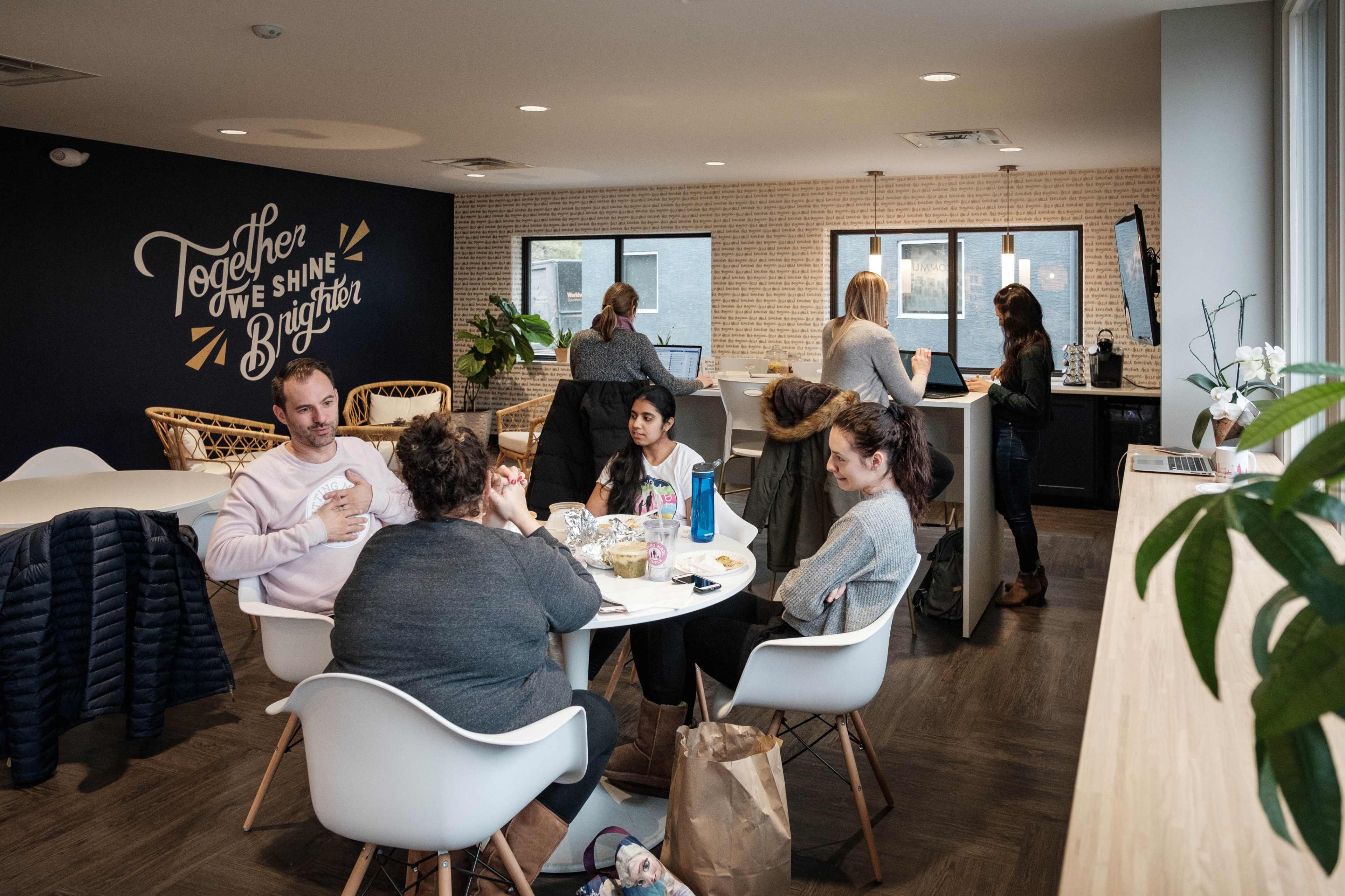 A group of people are seated at a round table in a modern workspace, discussing and working, while others are standing nearby at a counter.