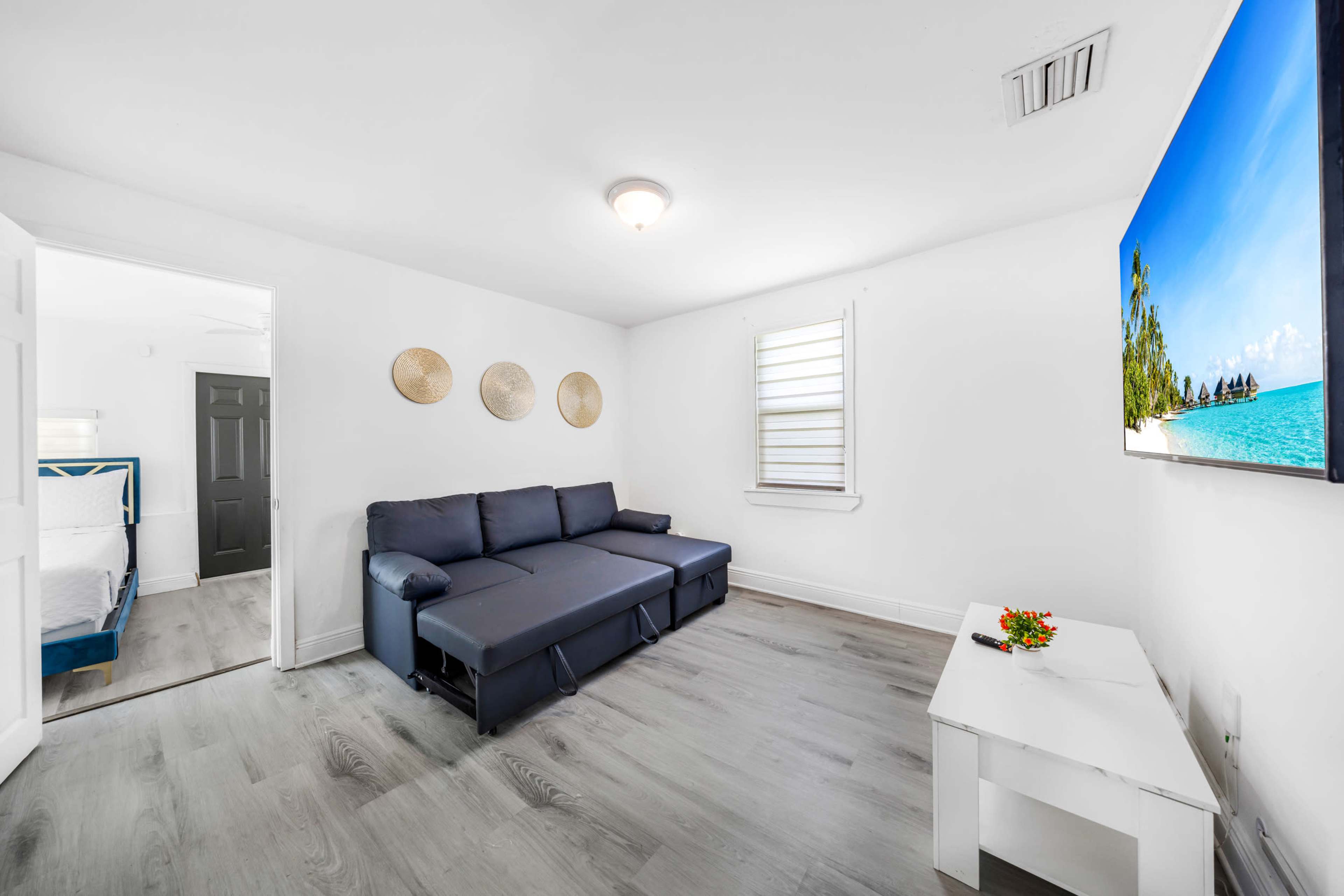 The image shows a minimalist living room with a dark sectional sofa, a white TV stand, and a wall-mounted television displaying a beach scene.