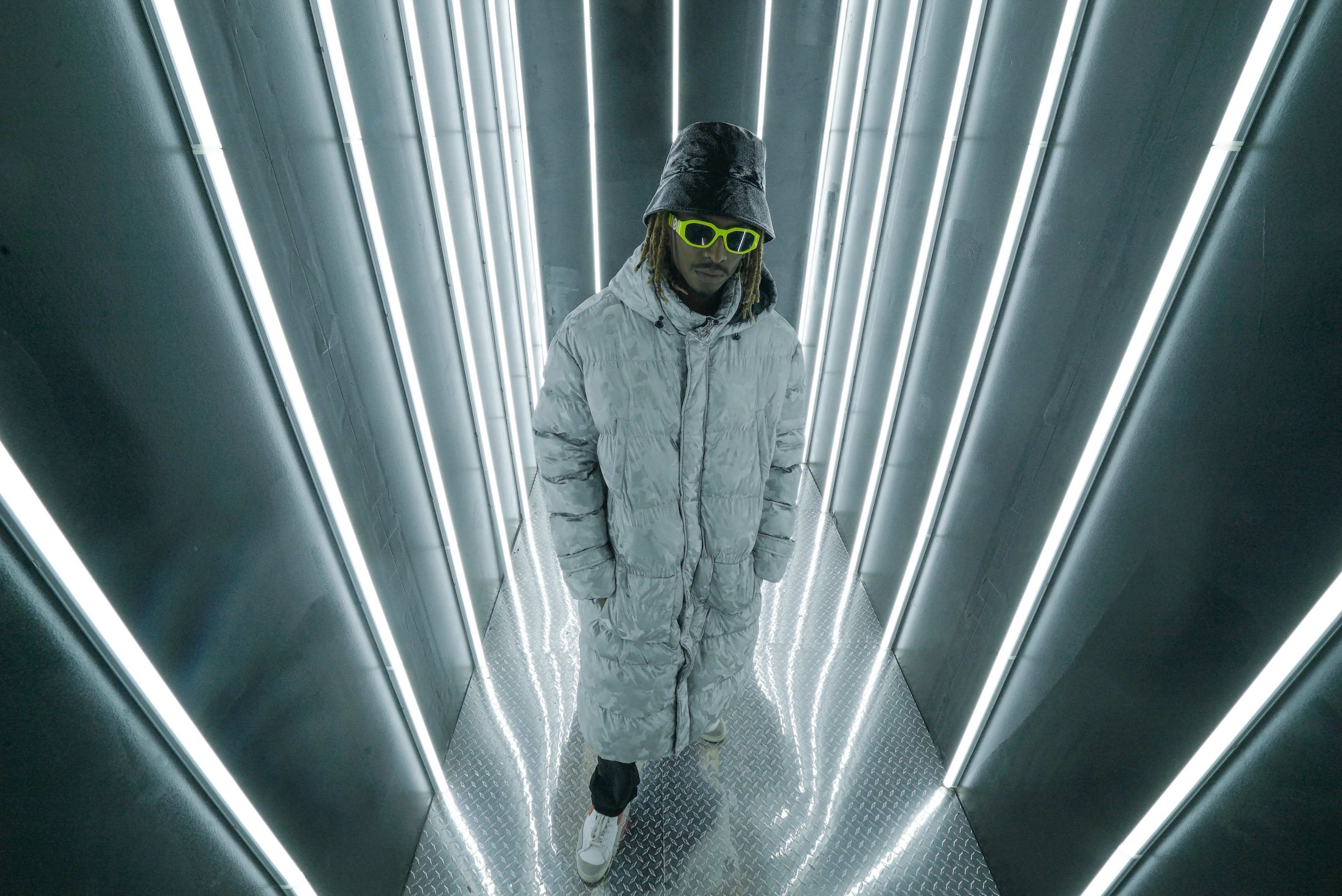 A person in a reflective, oversized jacket and bright sunglasses stands in a narrow, illuminated corridor lined with vertical lights.