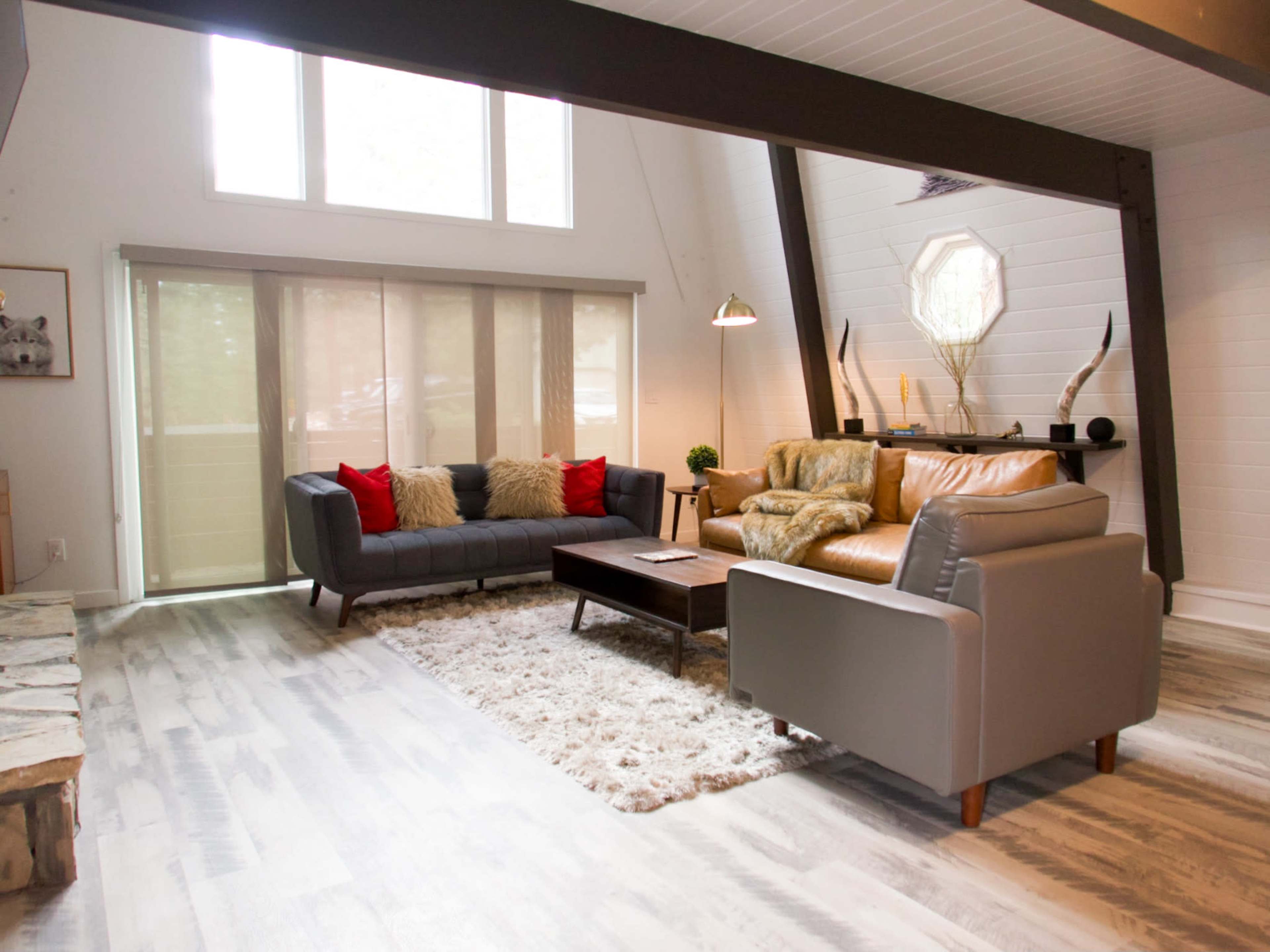 The living room features a mix of two sofas, a coffee table, and large windows, with wooden beams and a stone fireplace.