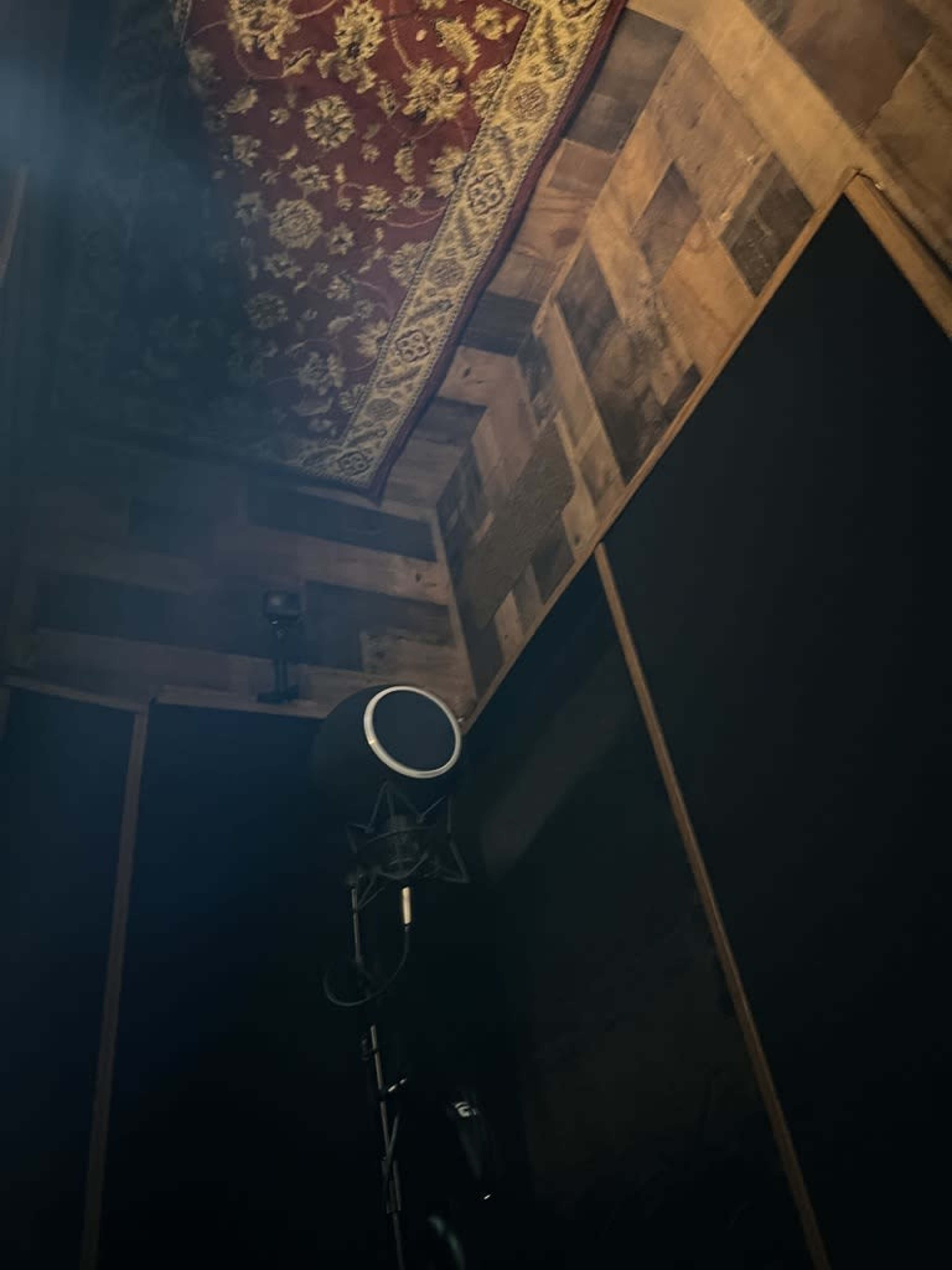 A recording booth with wooden walls, a microphone on a stand, and a patterned rug hanging from the ceiling.