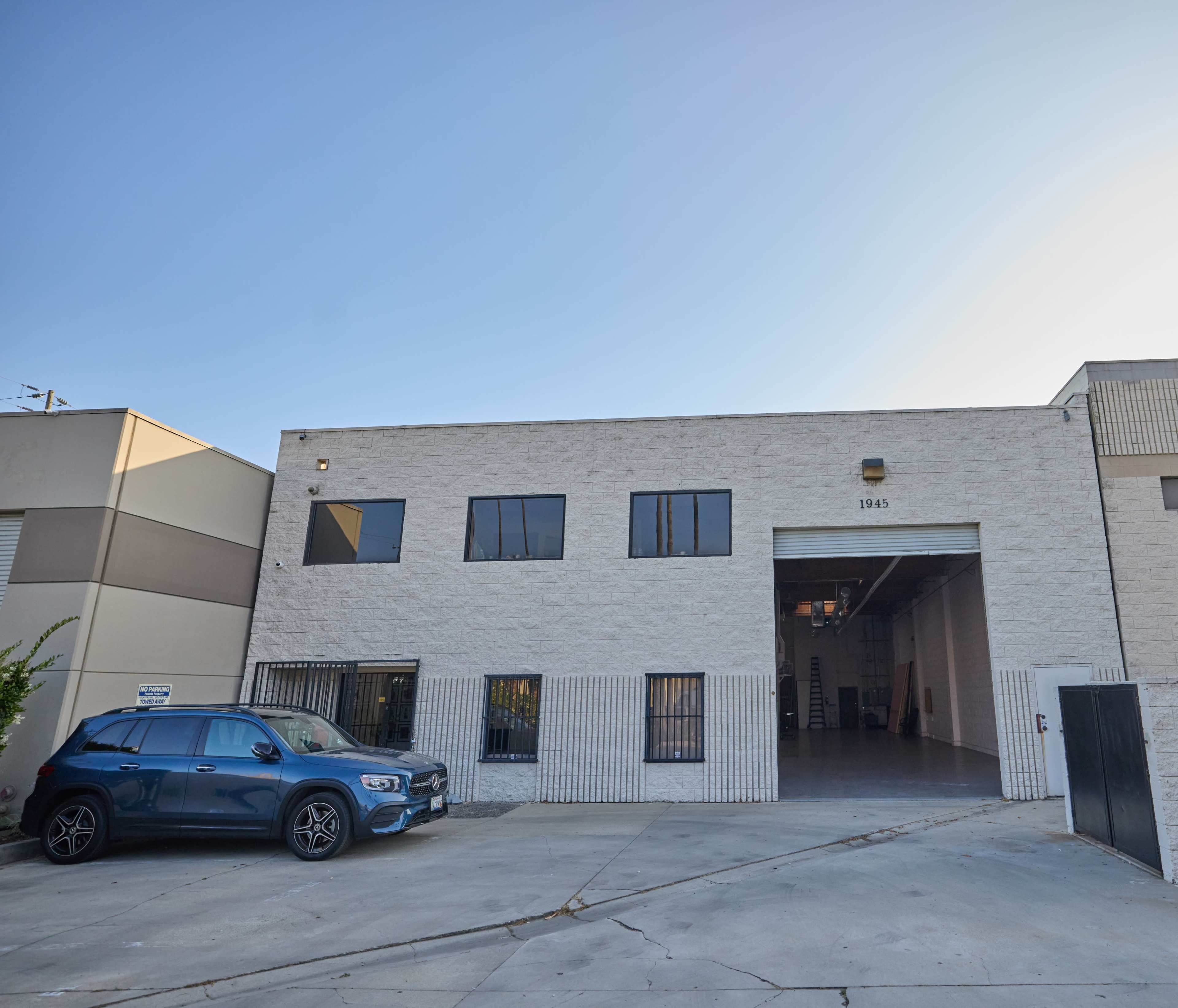 A white concrete building with large windows and an open garage area, flanked by two vehicles parked outside.