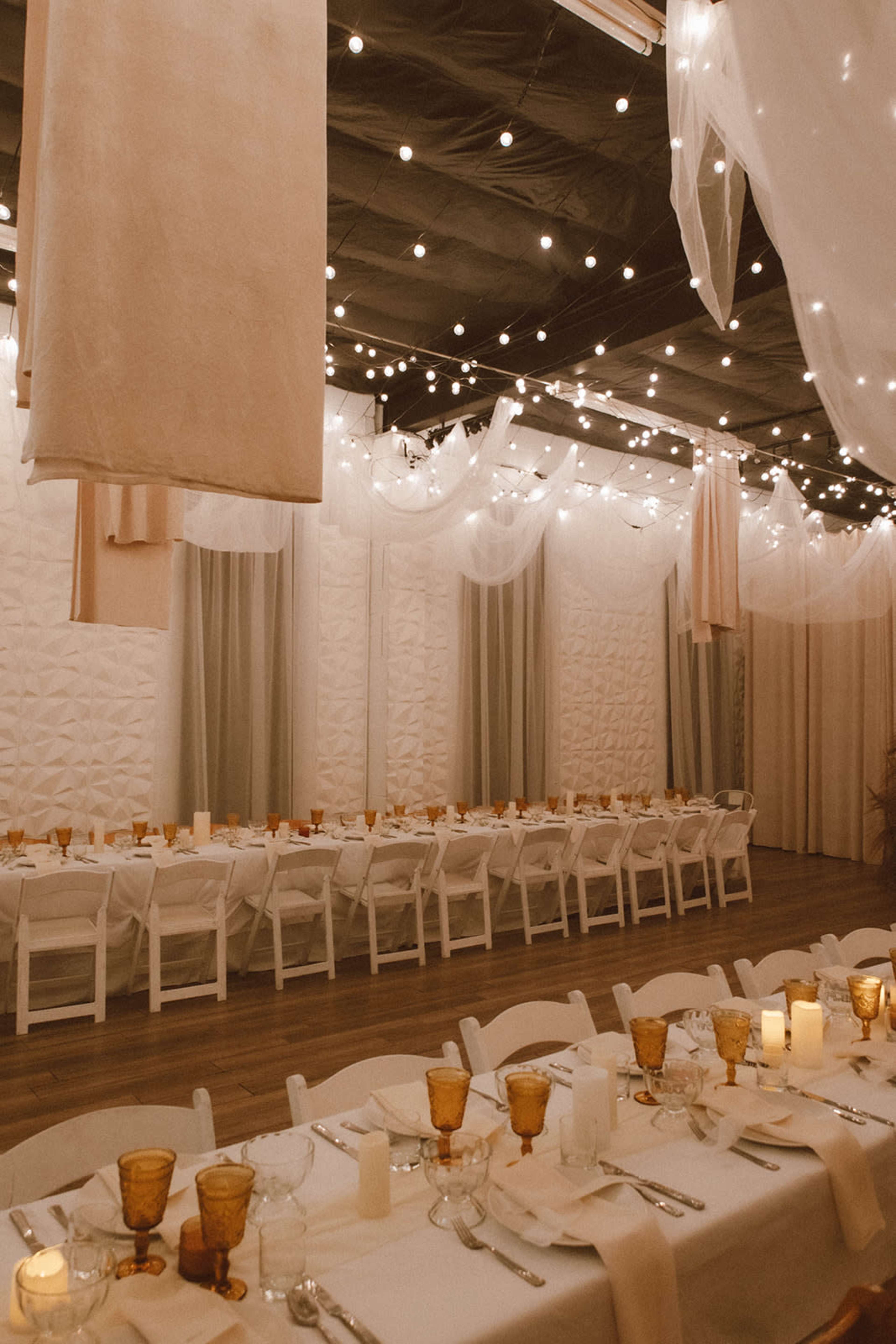 A banquet hall features long tables set with glasses and candles, beneath draped fabric and string lights.