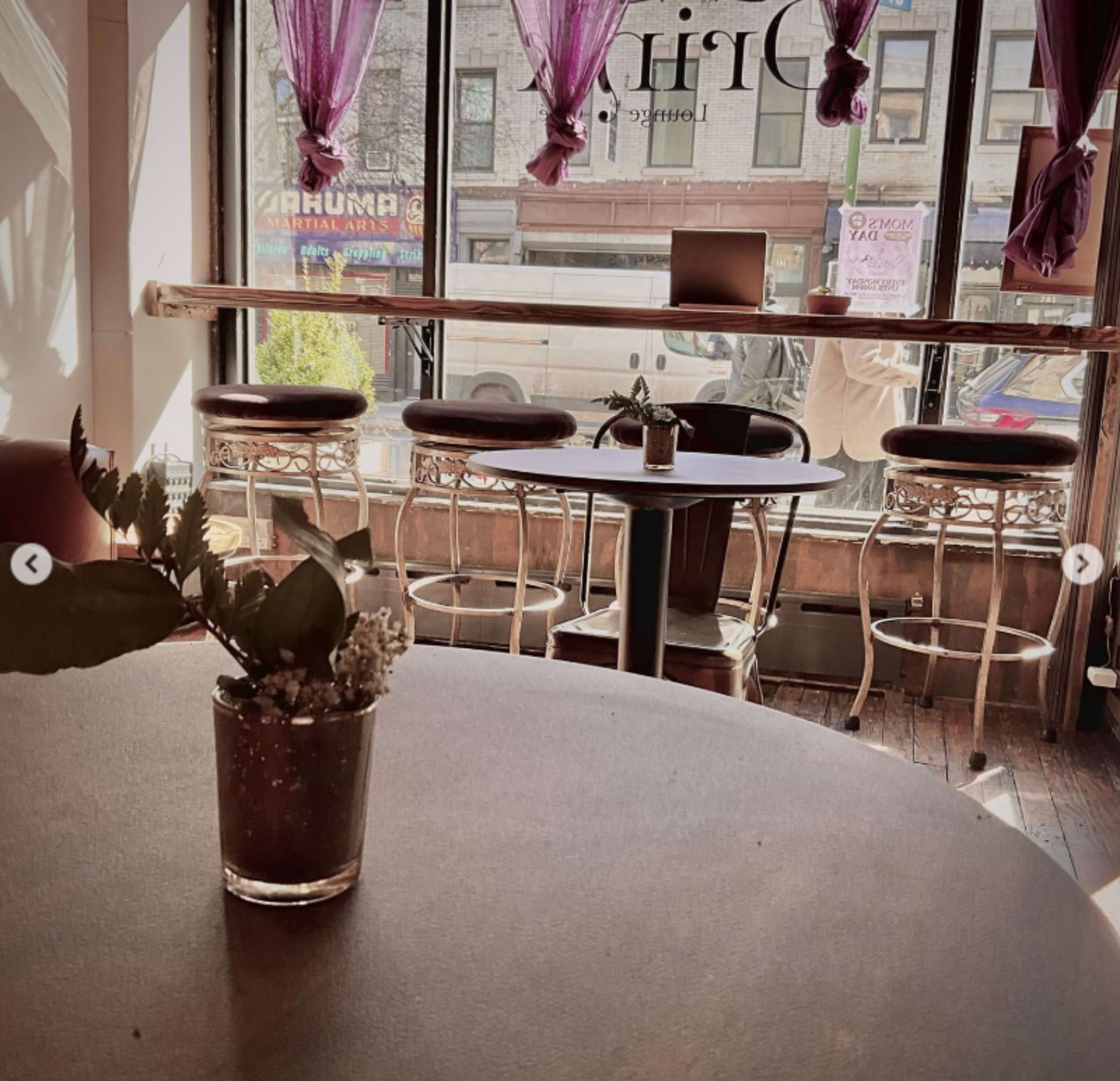 The image shows a cozy café interior with a wooden table, stools, and large windows draped with purple curtains.