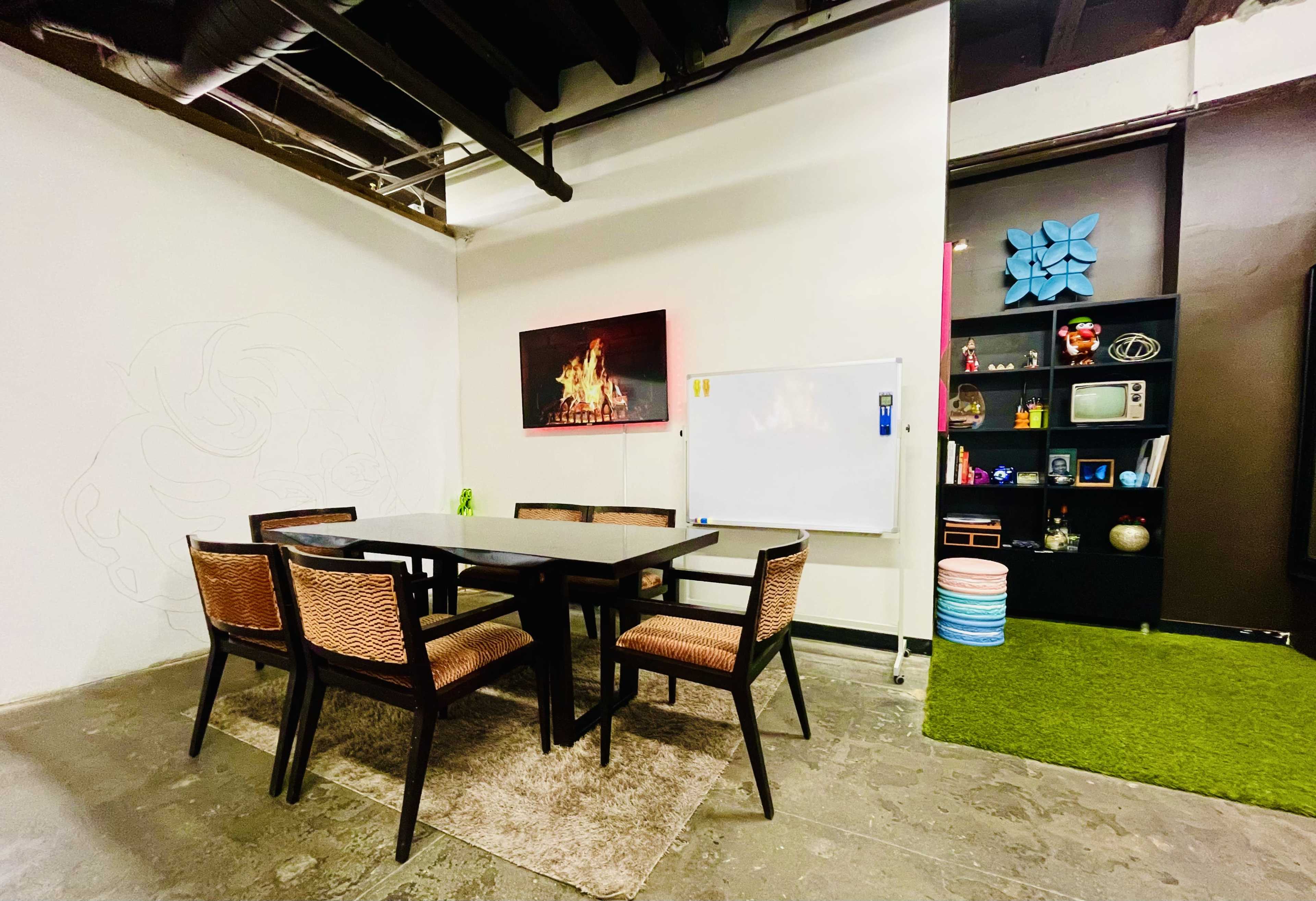 The image shows a modern meeting room with a rectangular table surrounded by chairs, a wall-mounted television displaying a fire animation, a whiteboard, and shelves with decorative items.