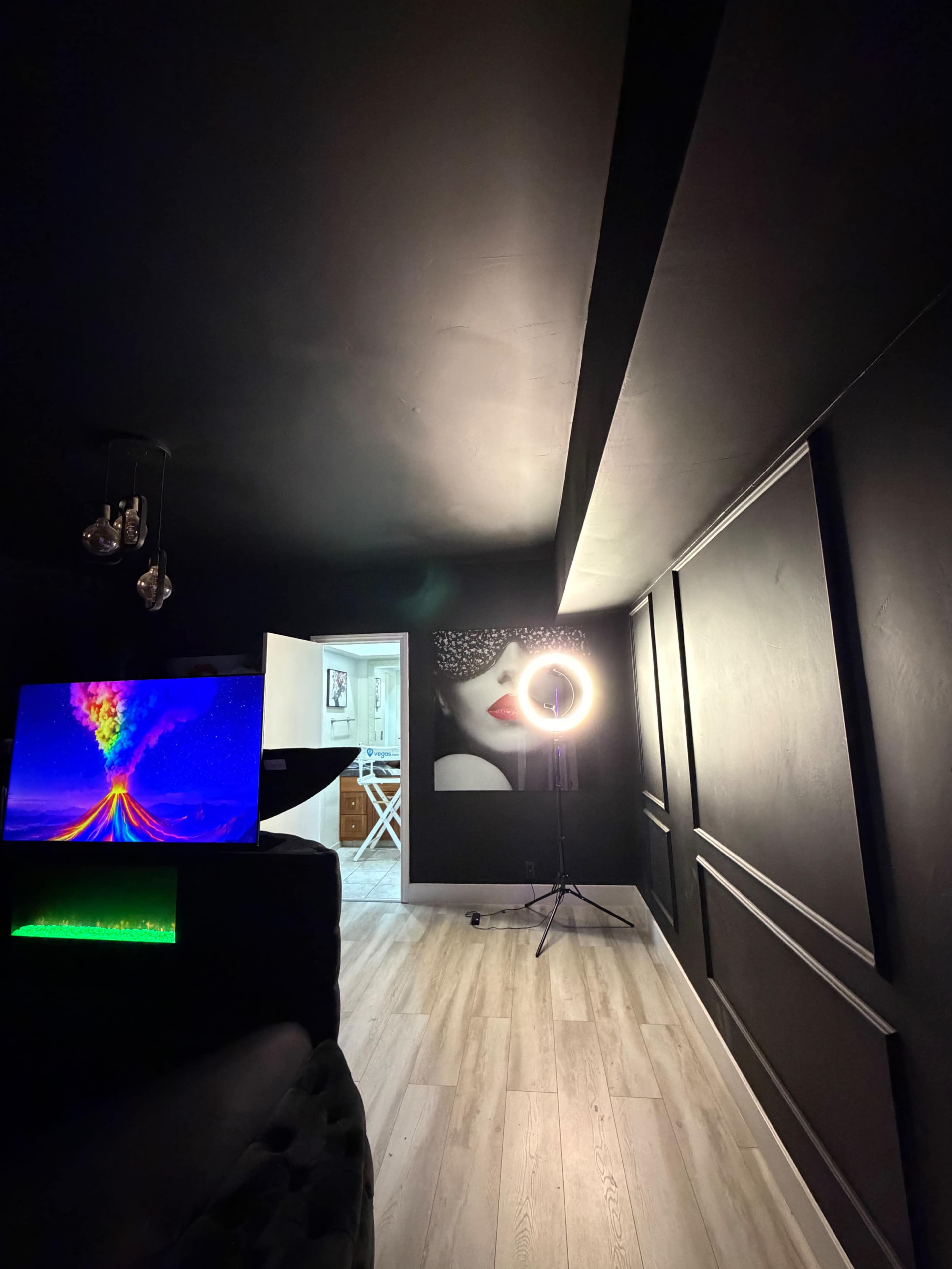 The image shows a dimly lit room with black walls, a large ring light on a stand, and a colorful visual display on a television.