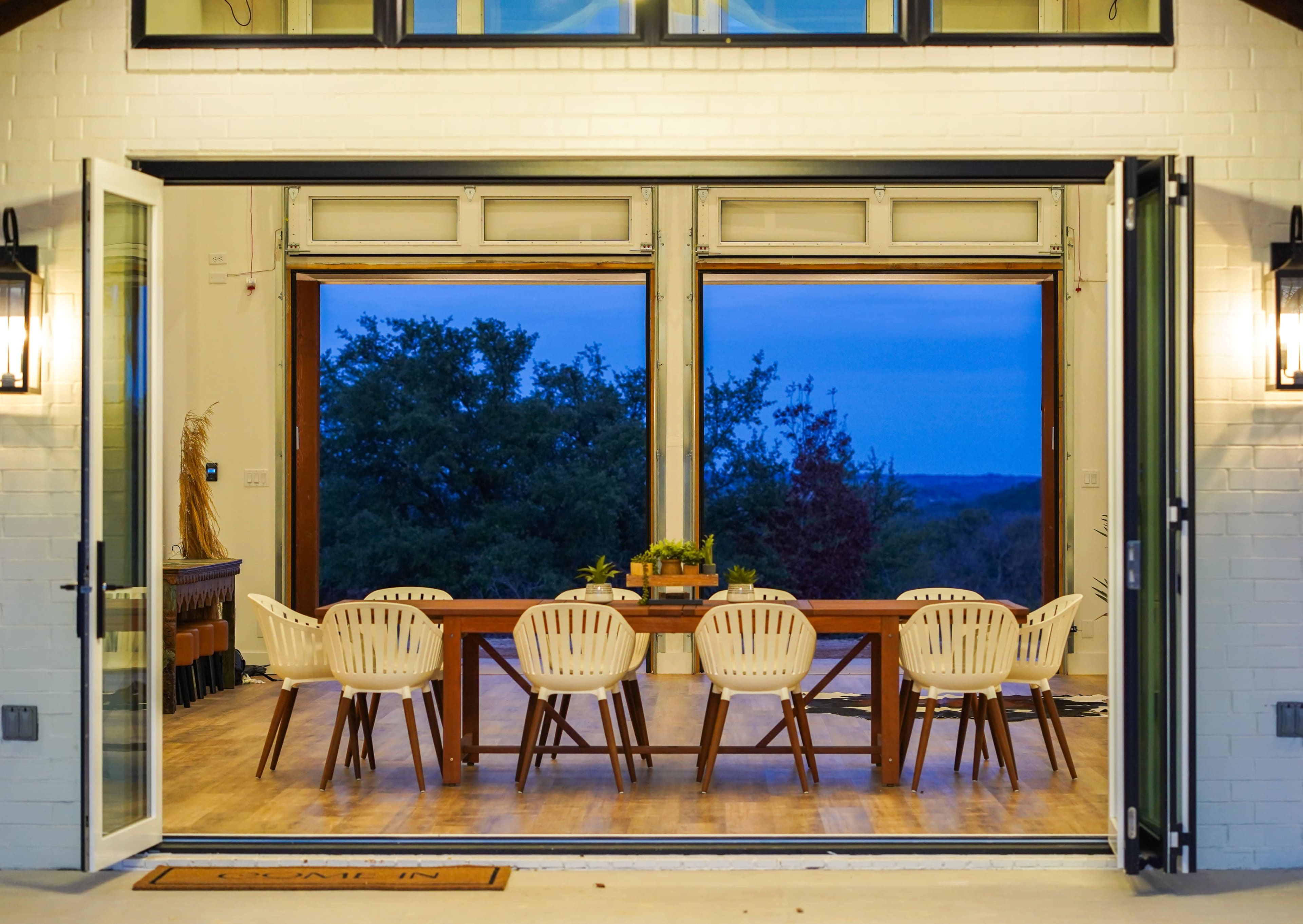 A wooden dining table with white chairs is positioned inside a spacious room that features large open doors, revealing a view of the landscape at dusk.