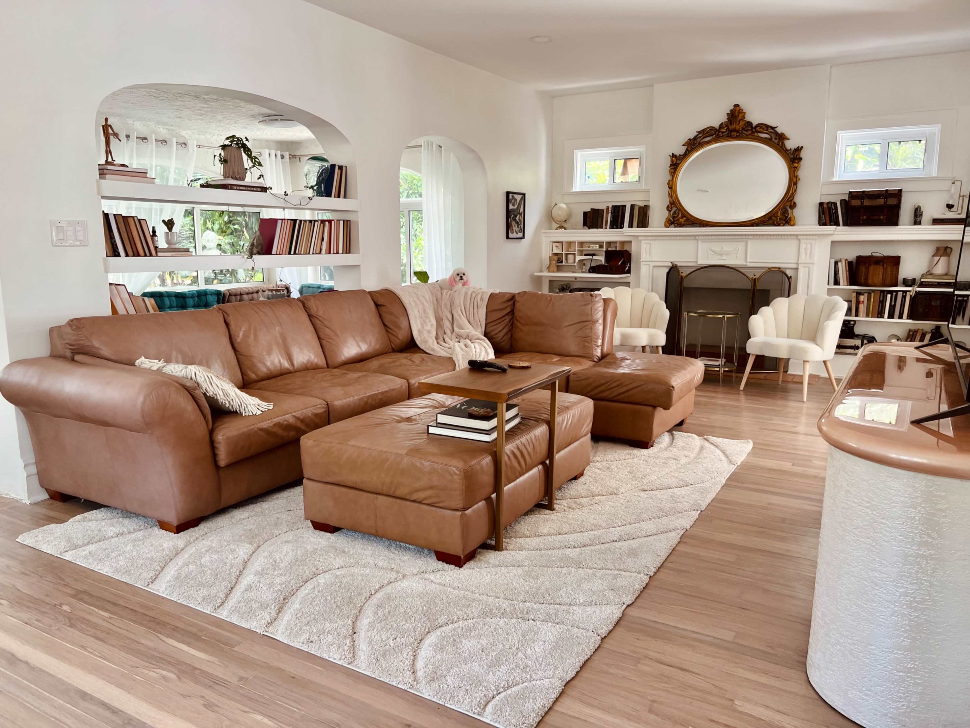 A spacious living room features a large brown leather sectional sofa, a central coffee table, and a light-colored area rug on wooden flooring.