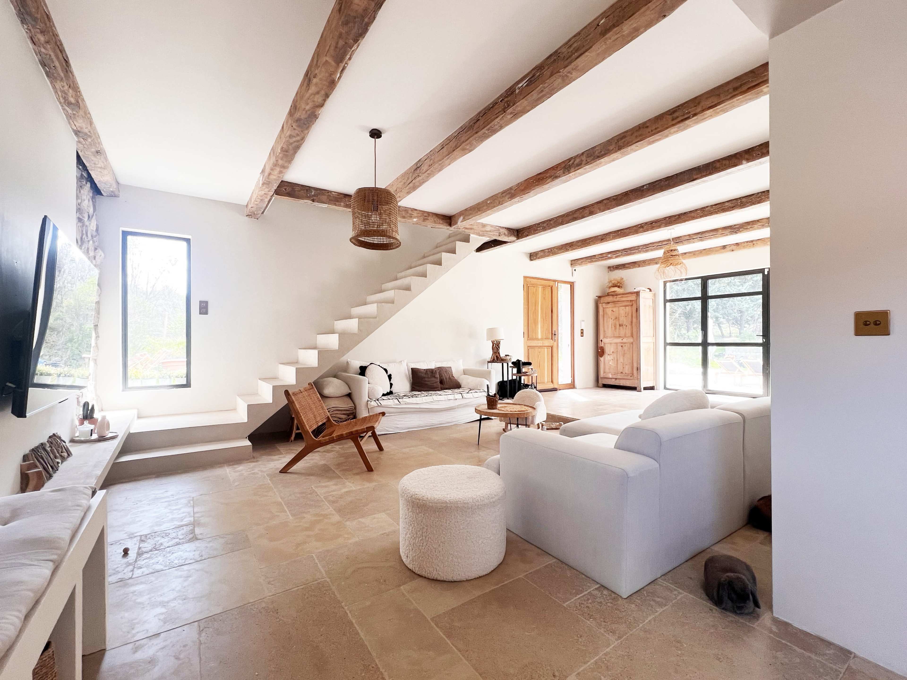 The image shows a spacious, modern living room with exposed wooden beams, a white sofa, a coffee table, and a staircase leading to an upper level.