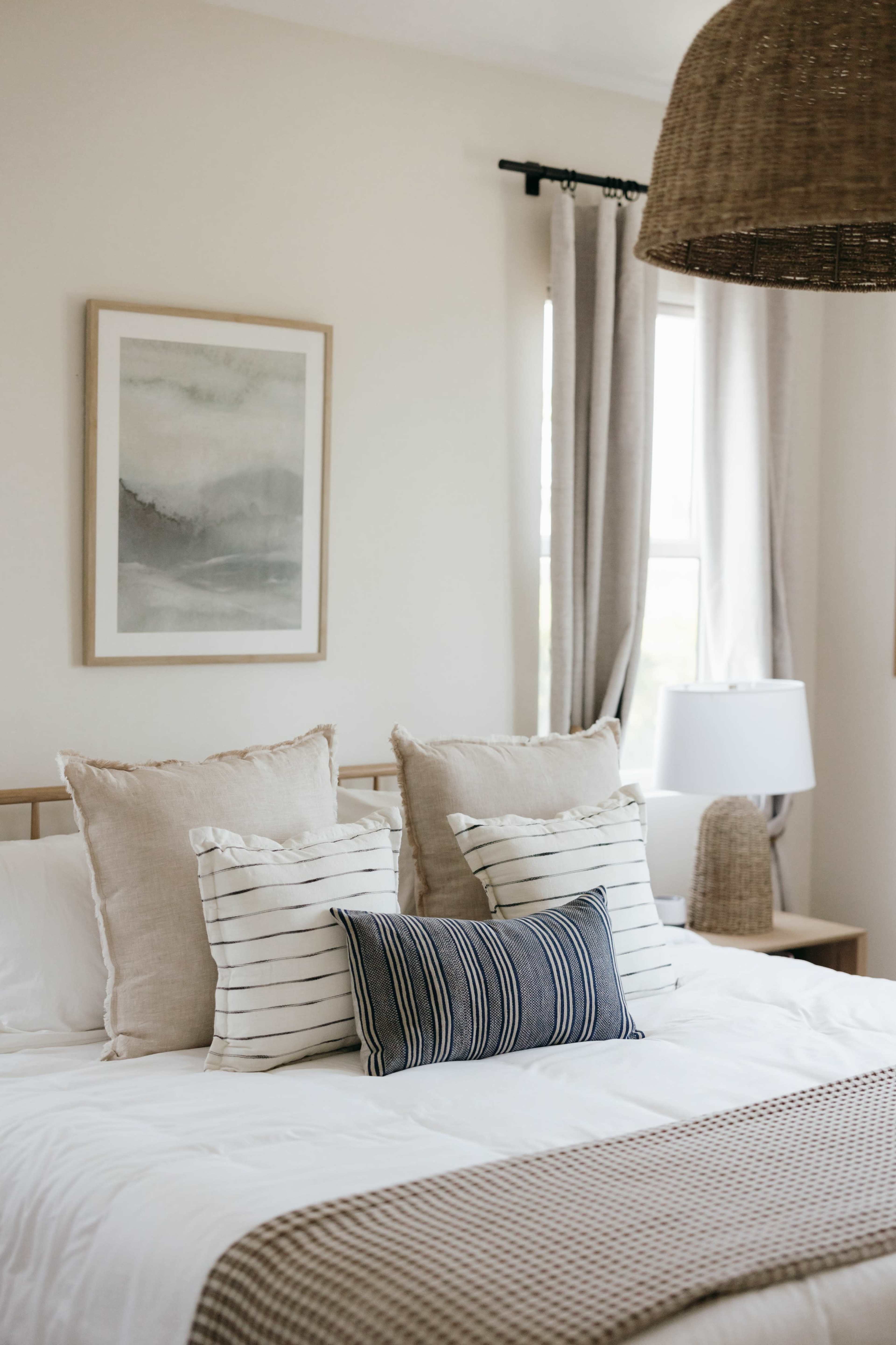 The image shows a neatly made bed with various decorative pillows, a framed artwork on the wall, and a bedside lamp in a softly lit bedroom.