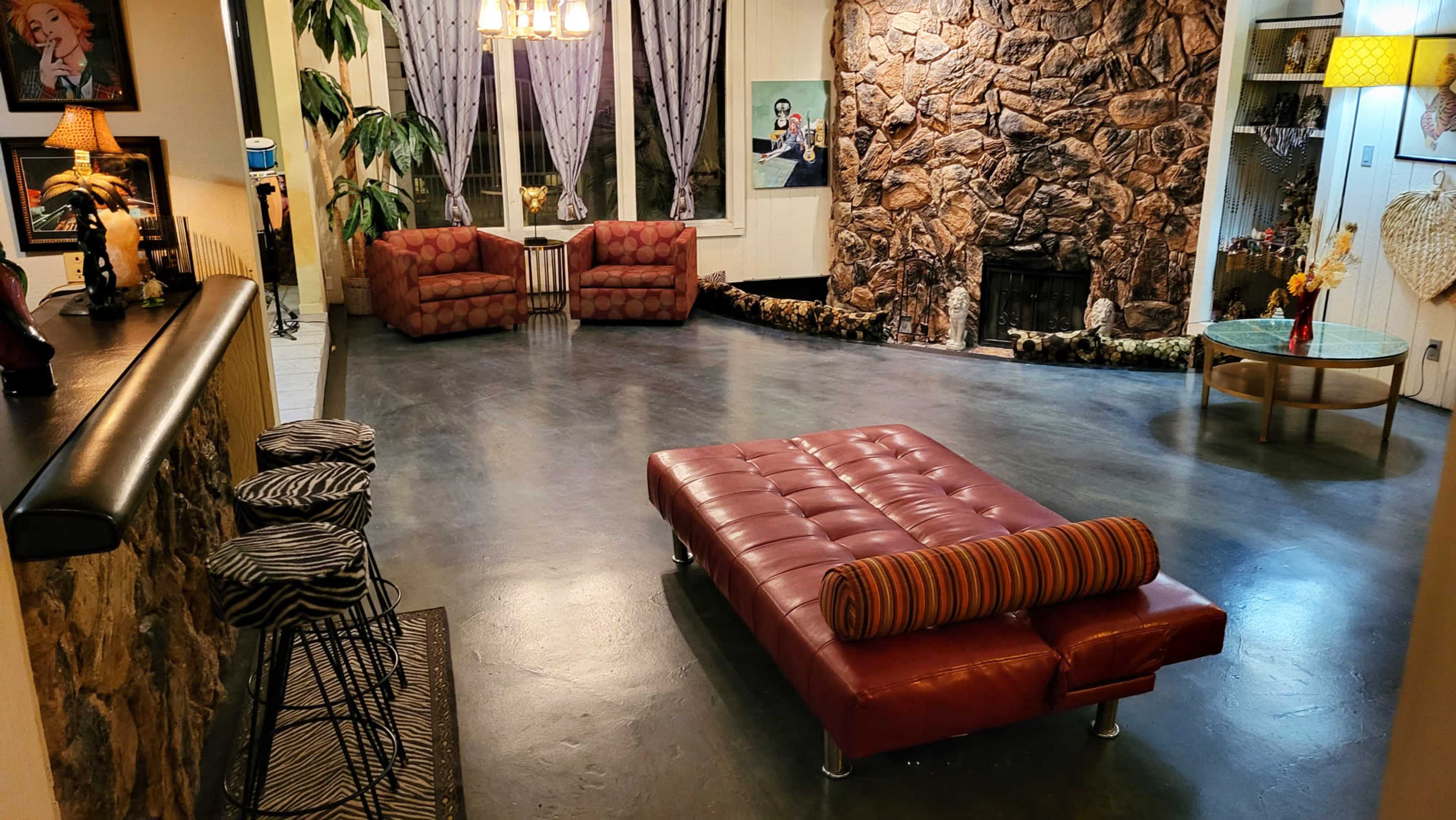 The image shows a spacious living room with two pink armchairs, a central red leather sofa, and a stone wall, complete with a fireplace and decorative plants.